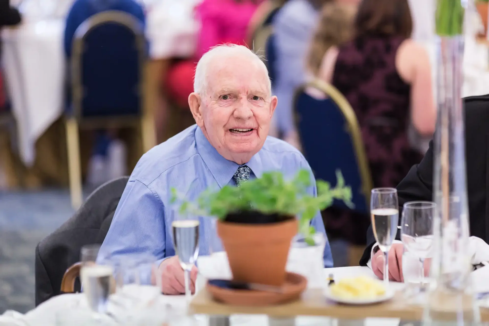 Elderly man in a blue shirt sitting at a banquet table, smiling, with a champagne glass and decorative plant in front of him.