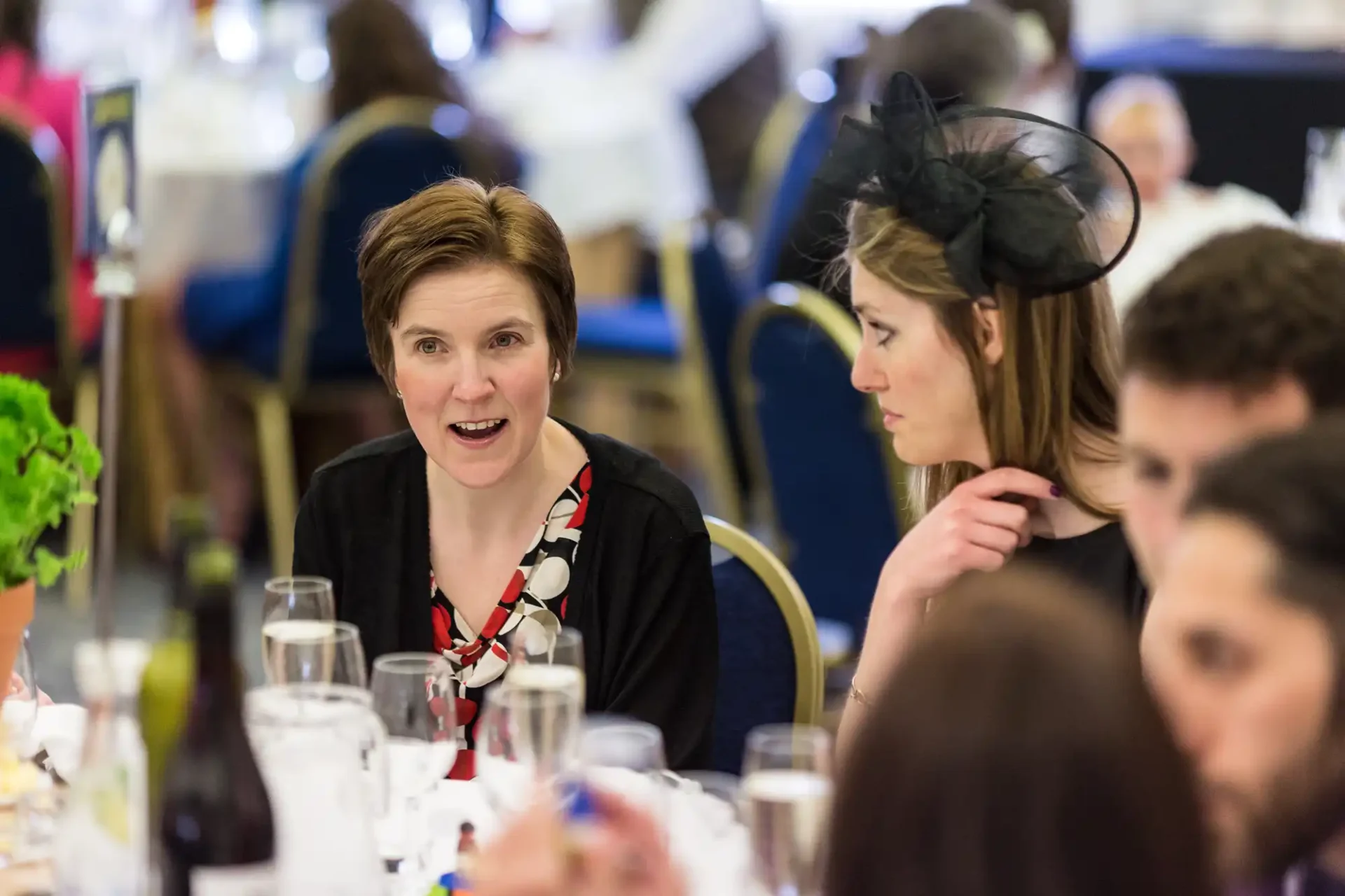 A woman in a black dress and red scarf, smiling at a formal dining event, while another woman in a black fascinator listens intently.