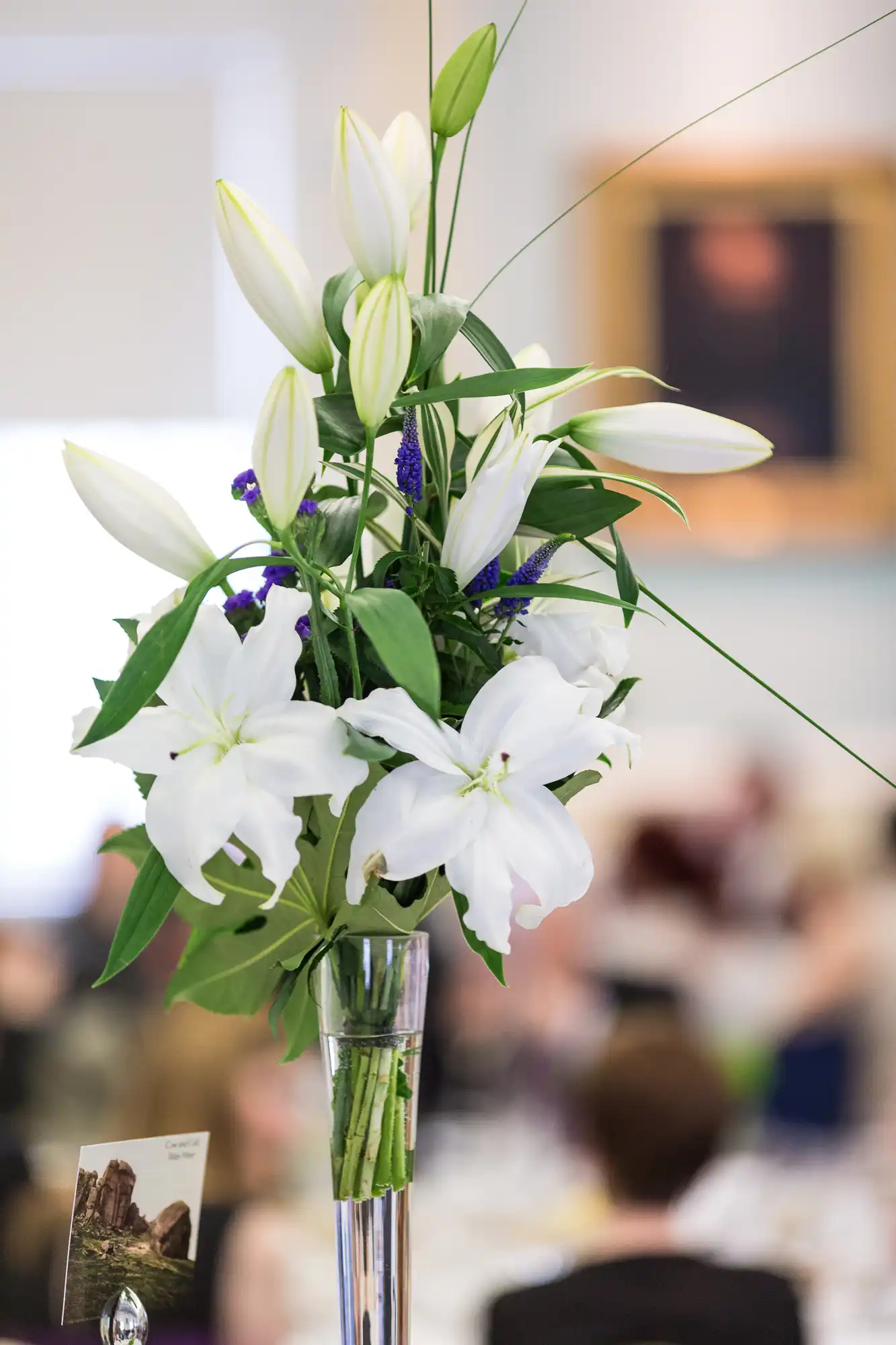 A tall vase filled with white lilies and purple flowers on a blurred background of a banquet hall setting.