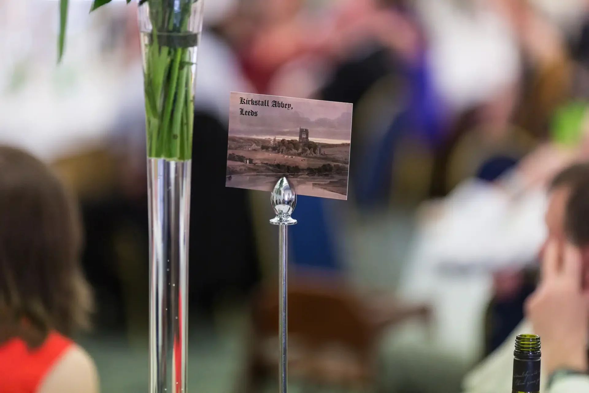 Postcard of birshtall abbey, kent, attached to a flower vase, in focus against a blurred background of people at an event.