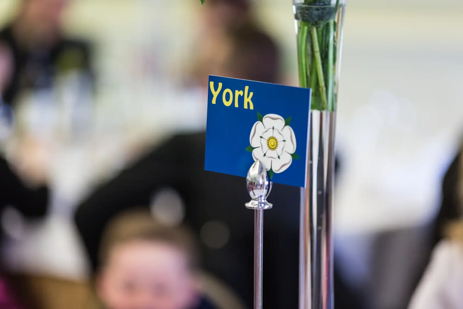 Table marker with "york" and a white flower emblem in a glass stand, with out-of-focus guests in the background.