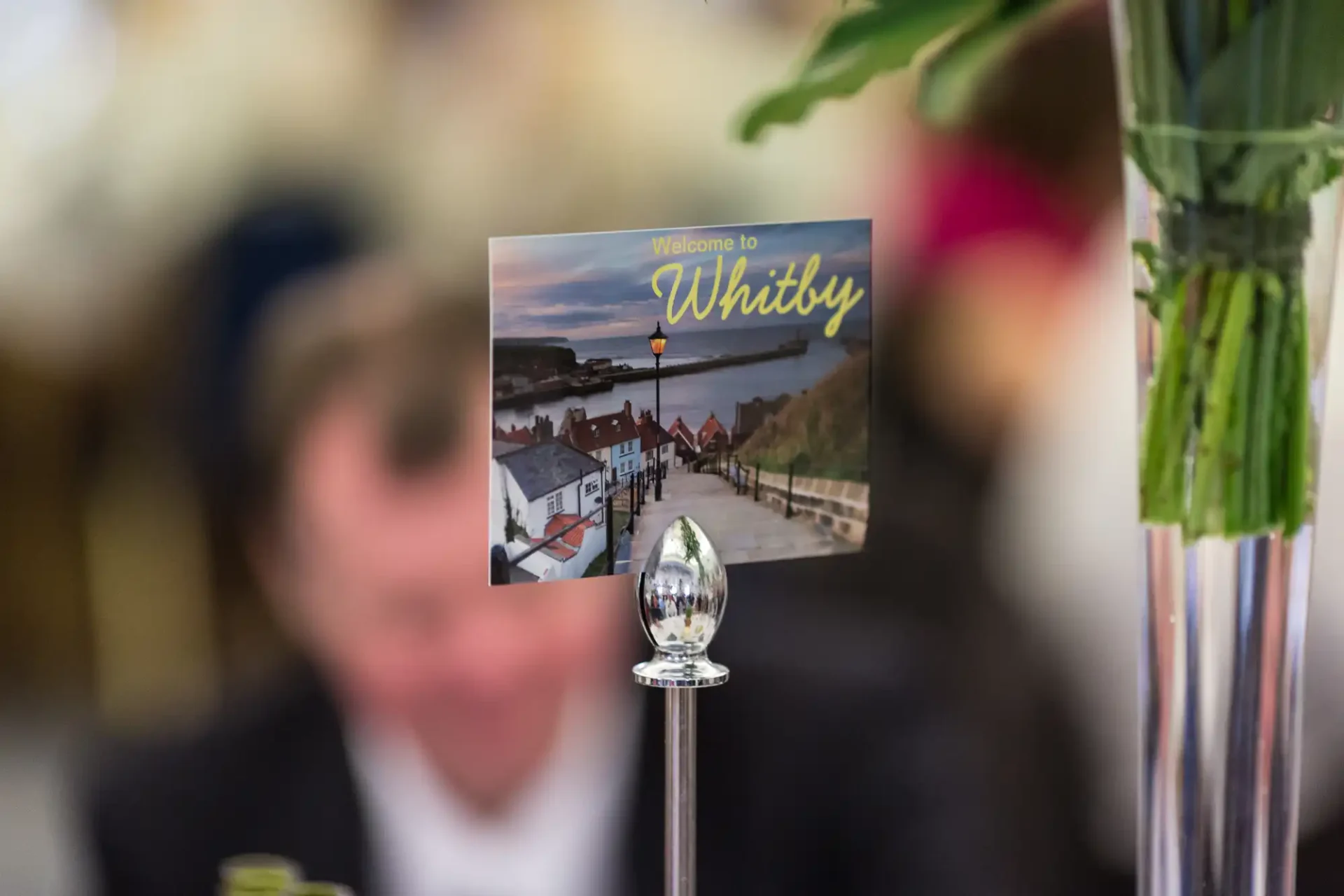 Table decoration featuring a clear drop-shaped glass with a blurred background showing a man and a "welcome to whitby" postcard.