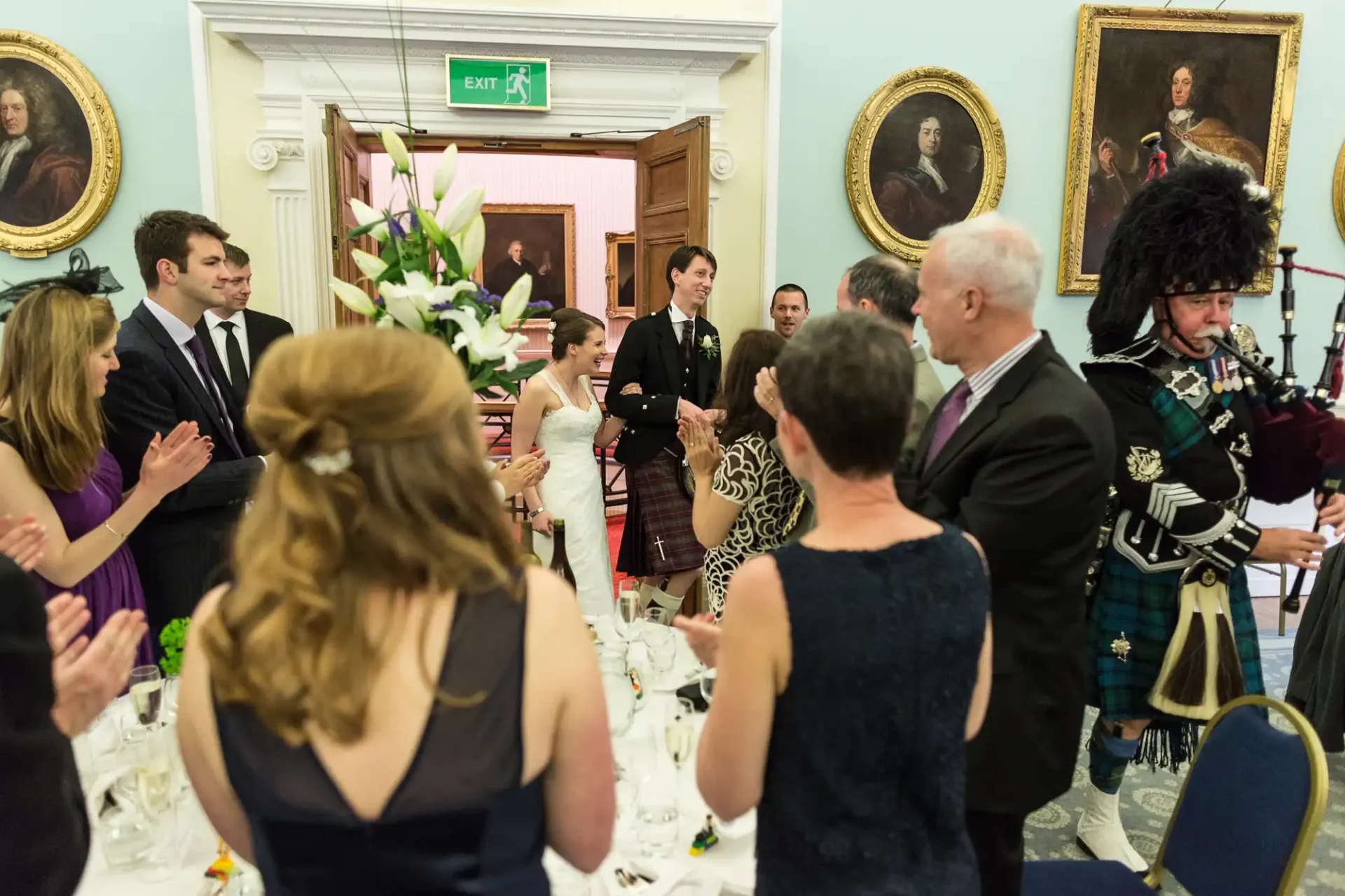 Guests clapping for a bride and groom at a wedding reception, with a bagpiper in traditional scottish attire playing in the foreground.