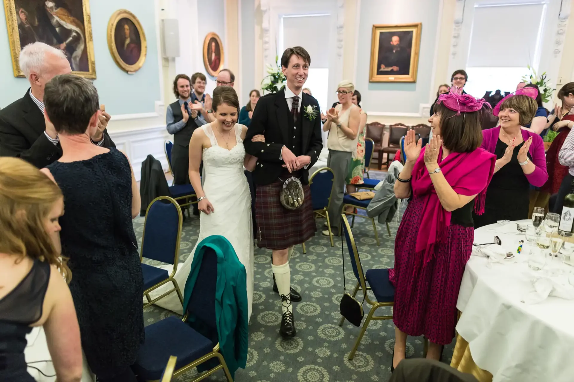 Bride and groom walking joyfully through a clapping crowd in a banquet hall, the groom dressed in a kilt and the bride in a white gown.