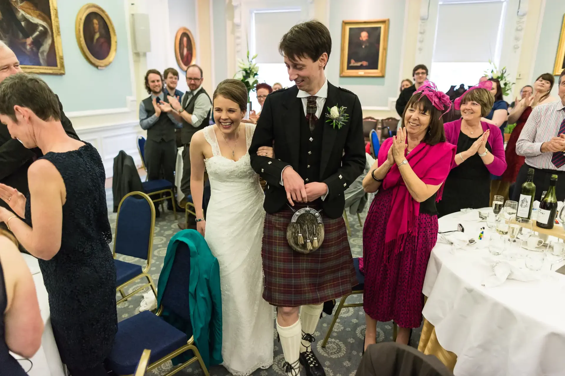 A bride and groom, the groom in a kilt, walking through applauding guests at their wedding reception.