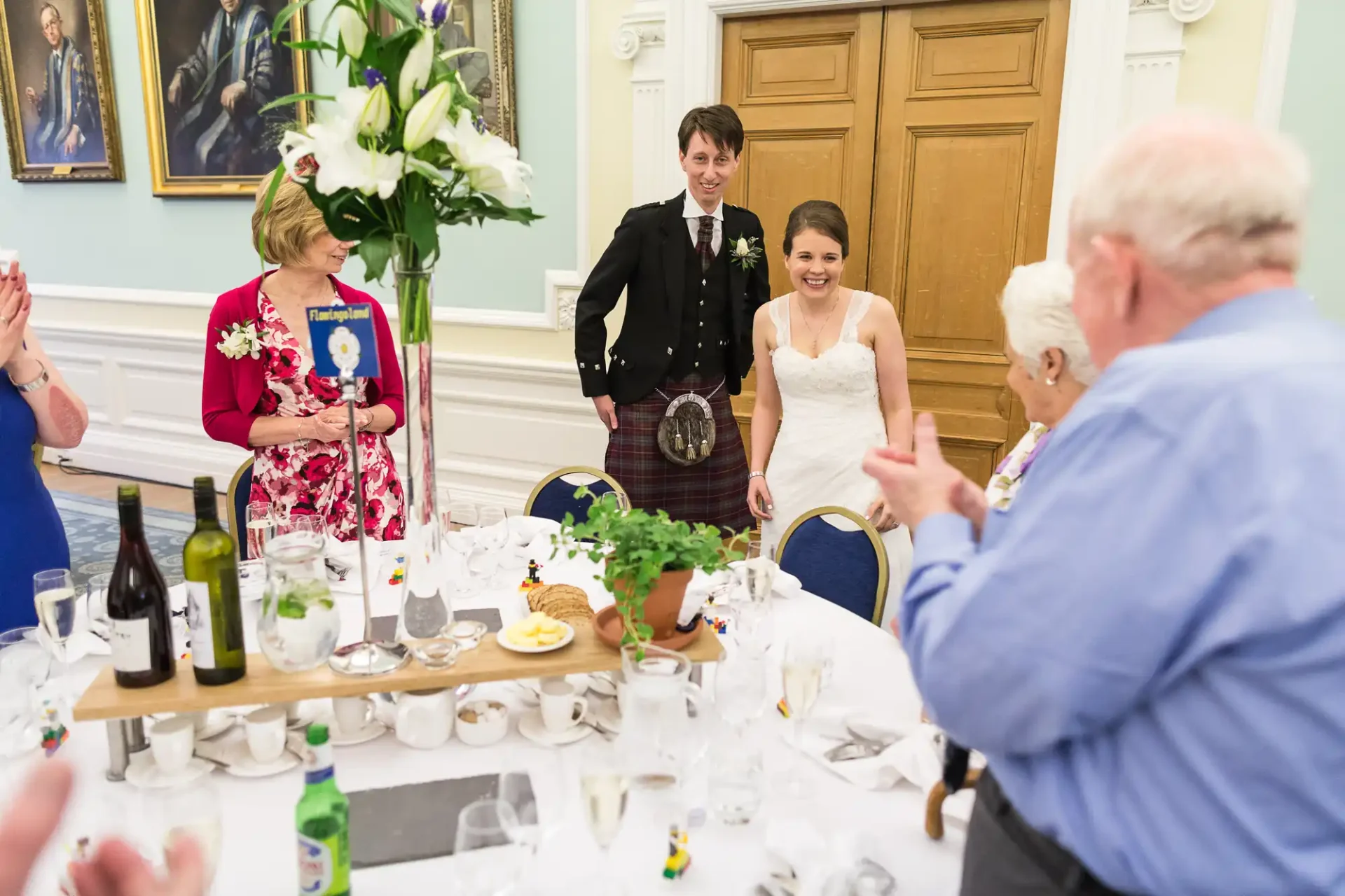 A newlywed couple smiles while walking through a clapping crowd at their wedding reception, the groom in a kilt and the bride in a strapless dress.