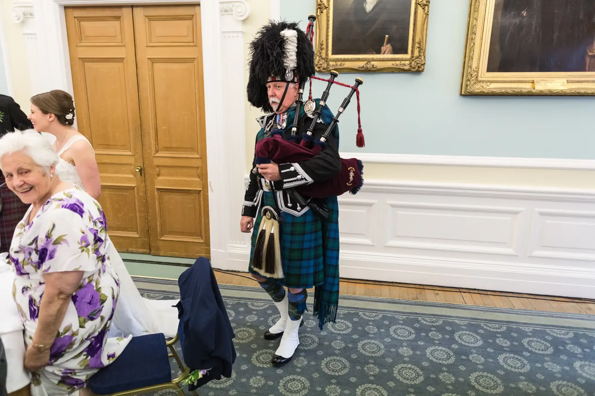 A man in traditional scottish attire, including a kilt and sporran, playing the bagpipes at an indoor event, walking past an elderly woman.