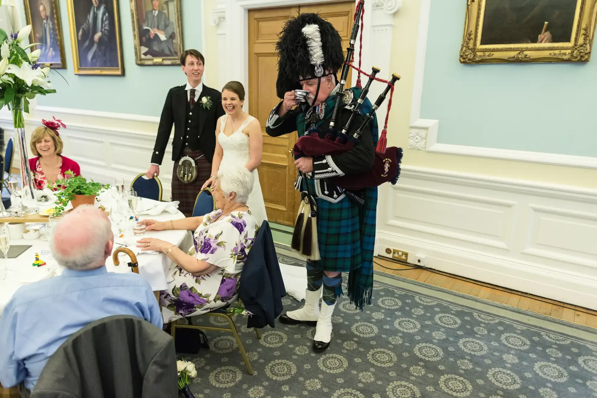 A bagpiper in traditional scottish attire plays at a wedding reception in an elegant room while a bride, groom, and guests watch and smile.