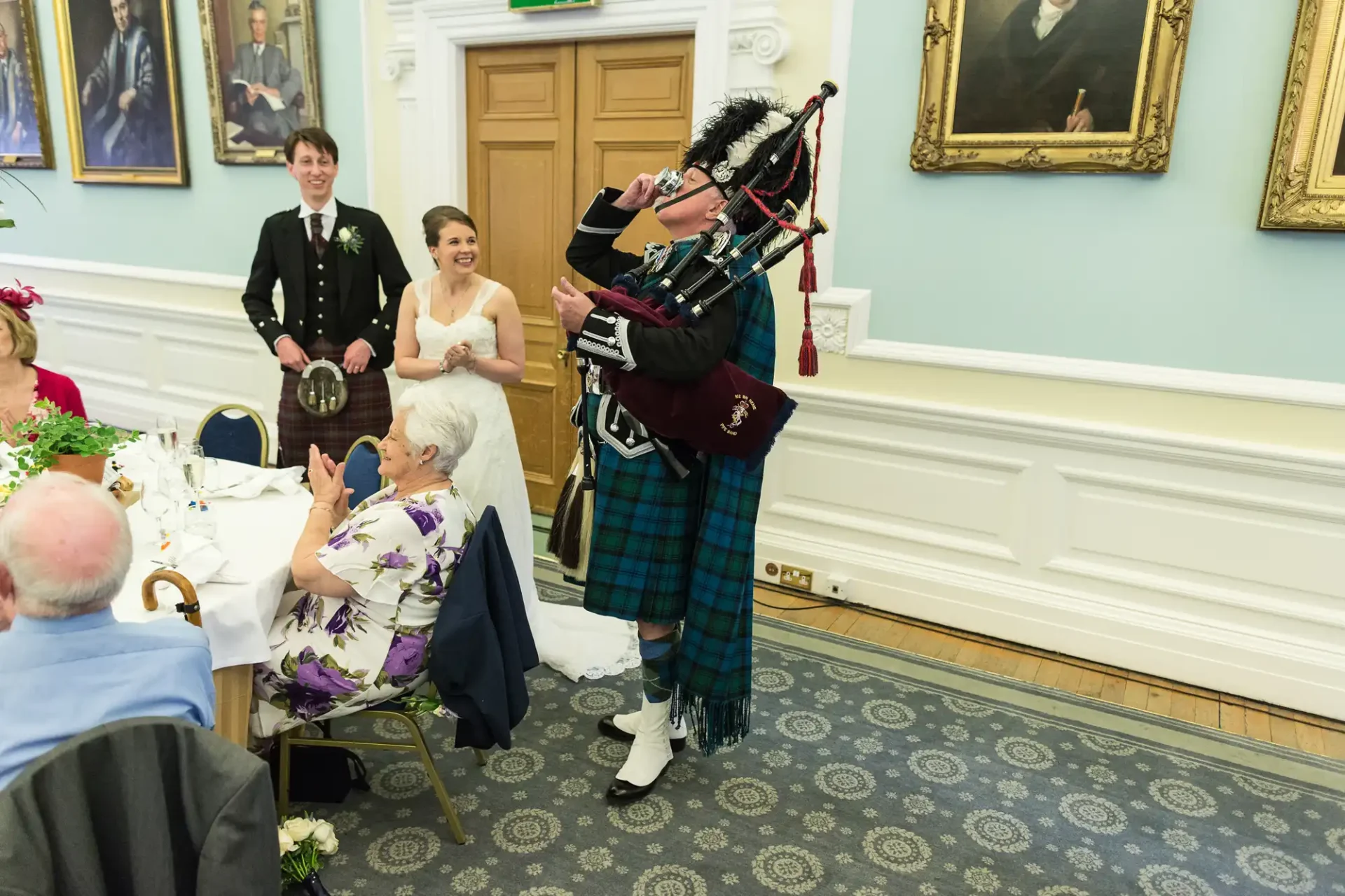 A bagpiper in traditional scottish attire playing at a wedding reception, with a smiling bride and groom seated in the background.