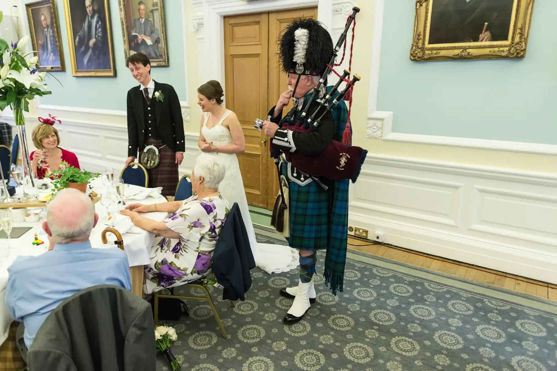 A bagpiper in traditional scottish attire playing at a wedding reception, with guests sitting around tables, observing and smiling.