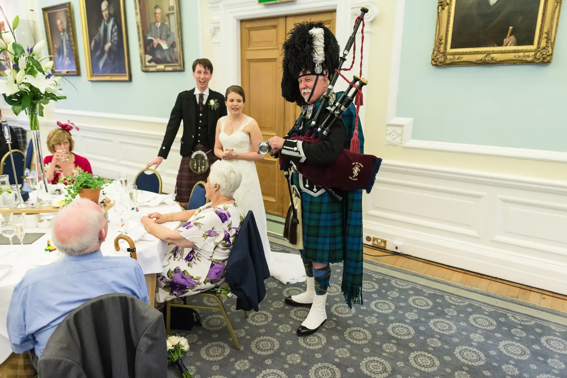 A bagpiper in traditional scottish attire plays at a wedding reception in an ornate room while guests watch.