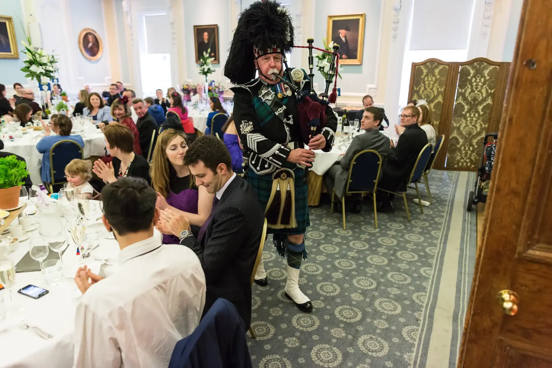 A bagpiper in traditional scottish attire playing at a formal indoor event with seated guests, some of whom are interacting warmly.