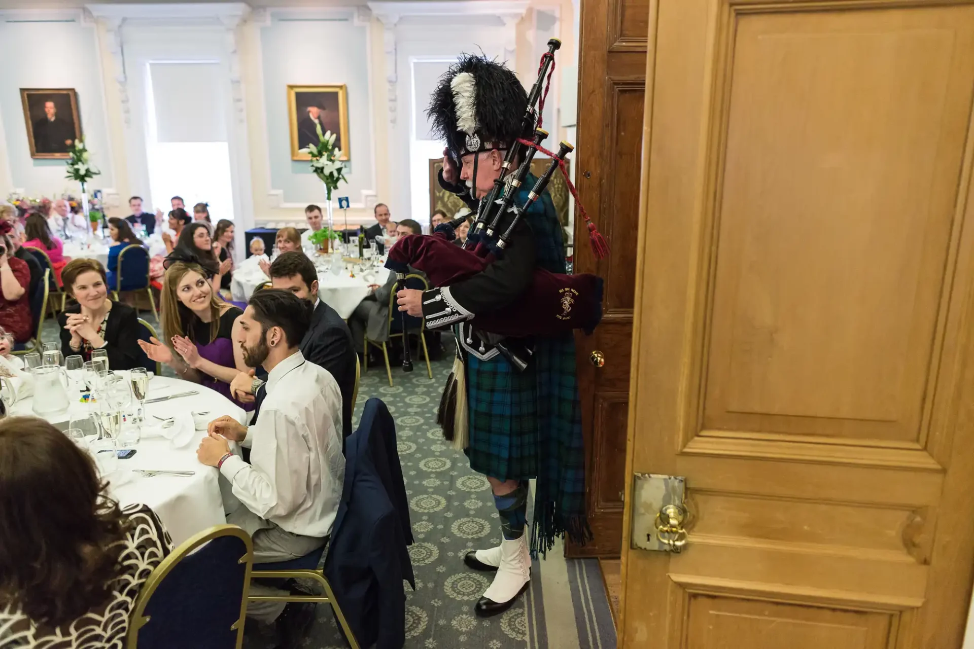 A bagpiper in traditional scottish attire playing for guests at a formal indoor dining event.