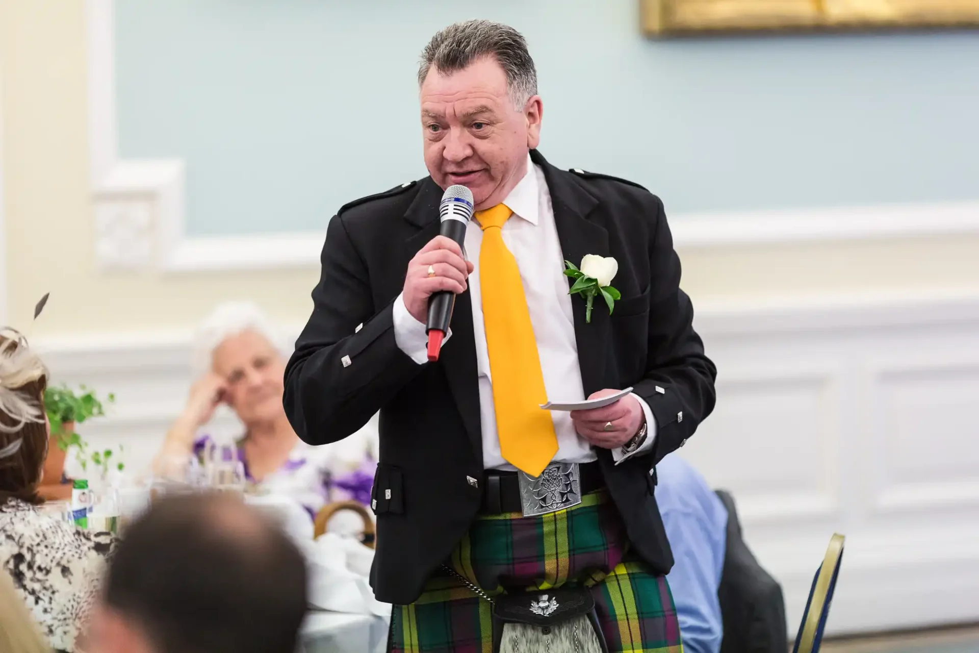 Man in kilt and tie speaking into a microphone at an event, holding a paper, with guests in the background.