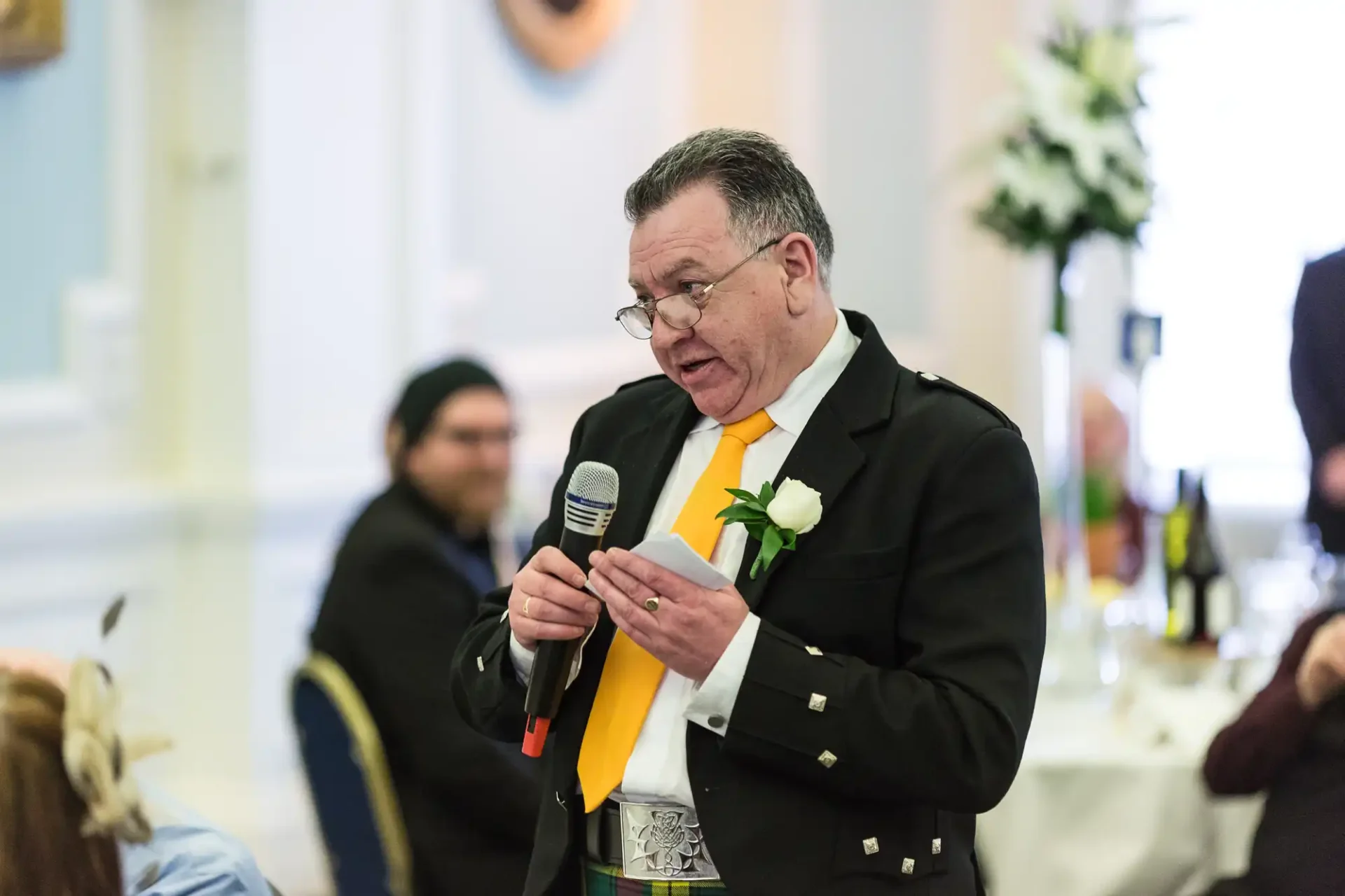 Man in black suit and yellow tie speaking into a microphone at a formal event, reading from a card, with blurred background.