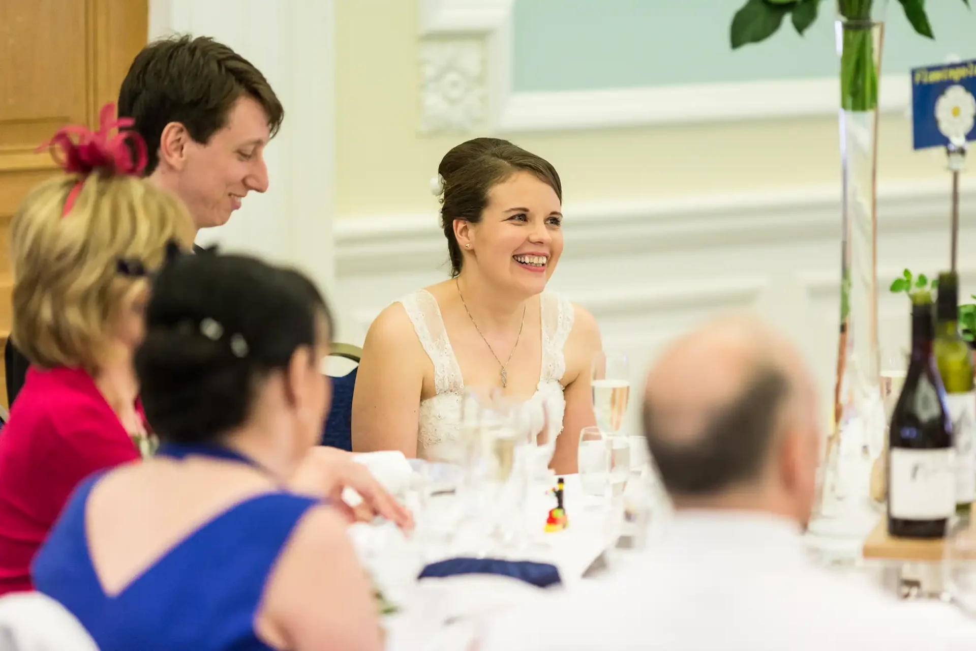 A bride in a white dress smiling at a wedding reception table surrounded by guests.