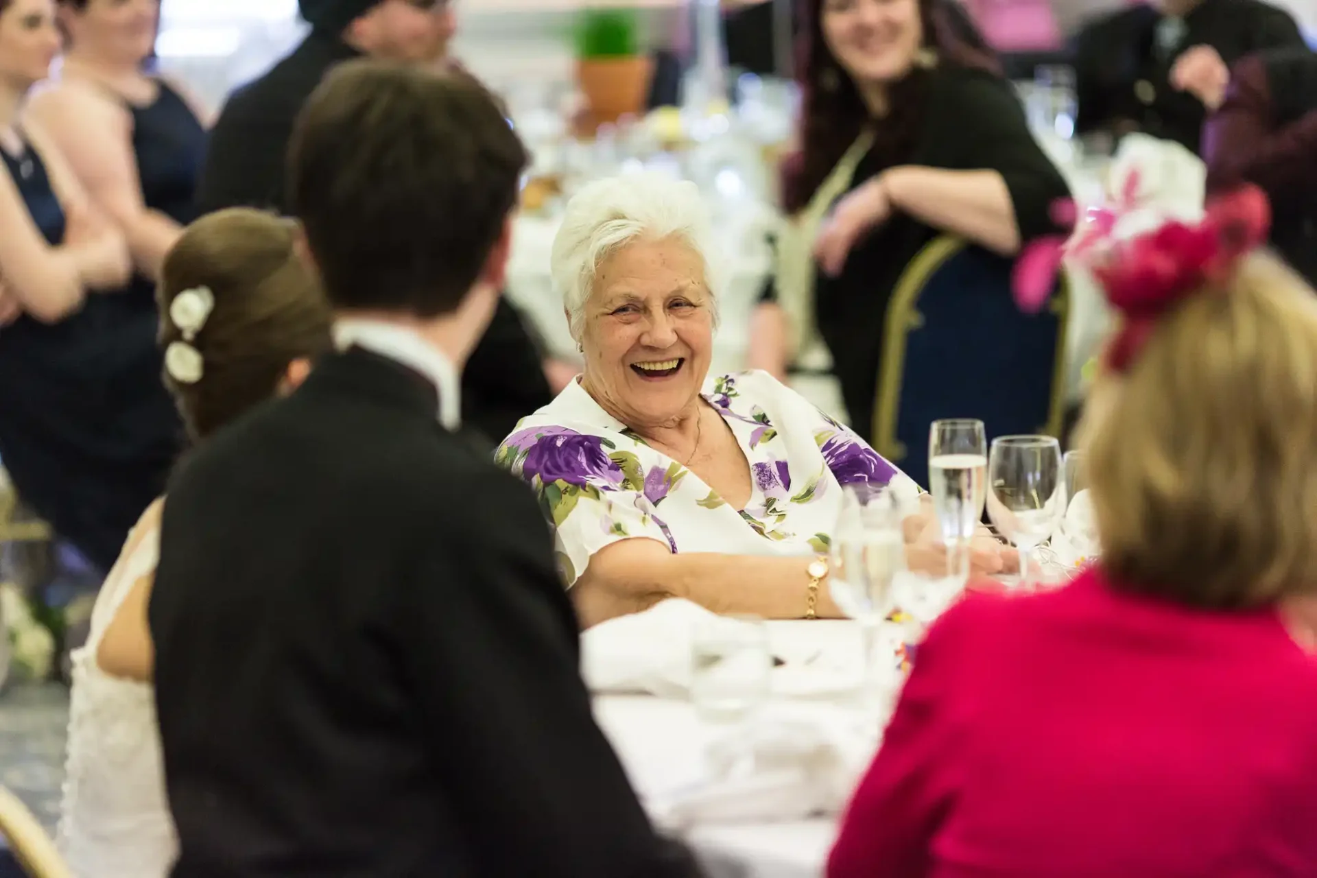 Elderly woman laughing joyfully at a wedding reception, surrounded by guests in a decorated venue.