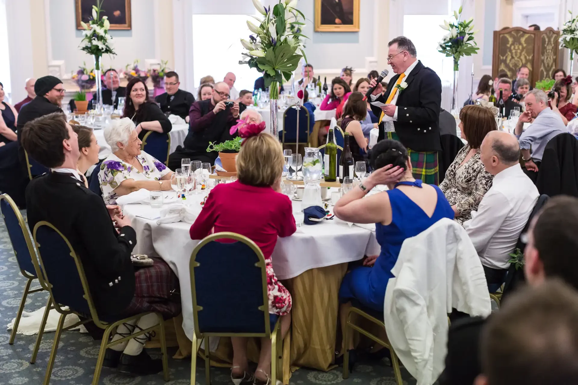 Man in kilt speaking to guests at a formal dinner event with tables set elegantly, guests engaged and dining in a brightly lit room.