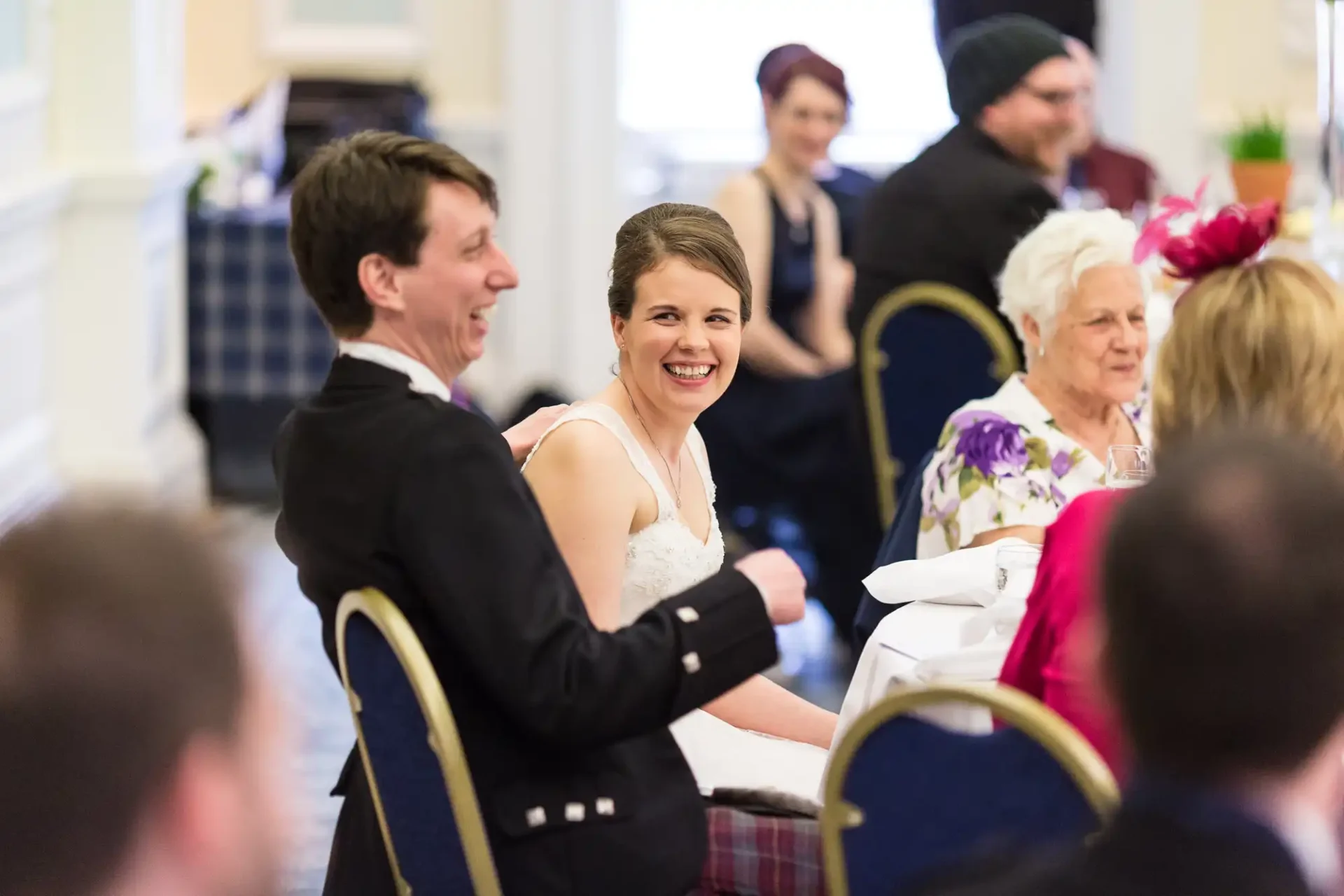 A bride and groom laugh joyfully at a wedding reception table, surrounded by guests in a brightly lit room.