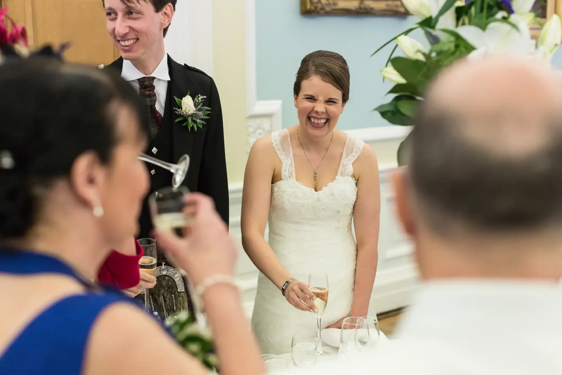 Bride laughing joyfully at a table during a wedding reception, with a groom next to her and guests interacting around them.