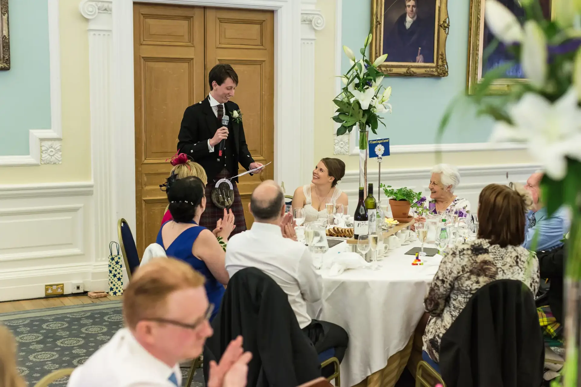 A man in a tuxedo giving a speech at a wedding reception, with guests seated at tables listening and smiling, in an elegant room with white lilies on the tables.