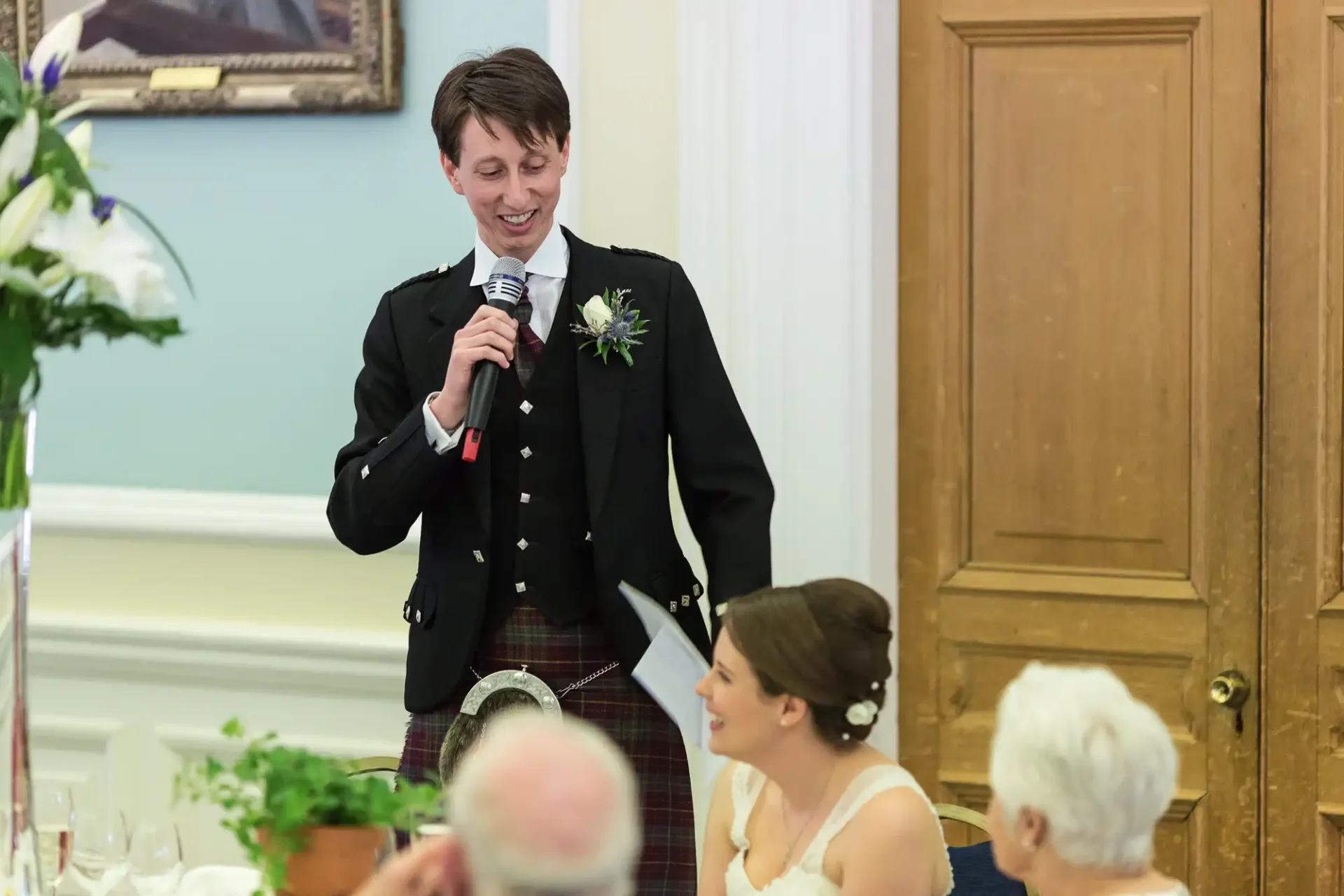 Man in a kilt and jacket speaking into a microphone at a wedding reception, smiling at a seated woman who is looking at him.