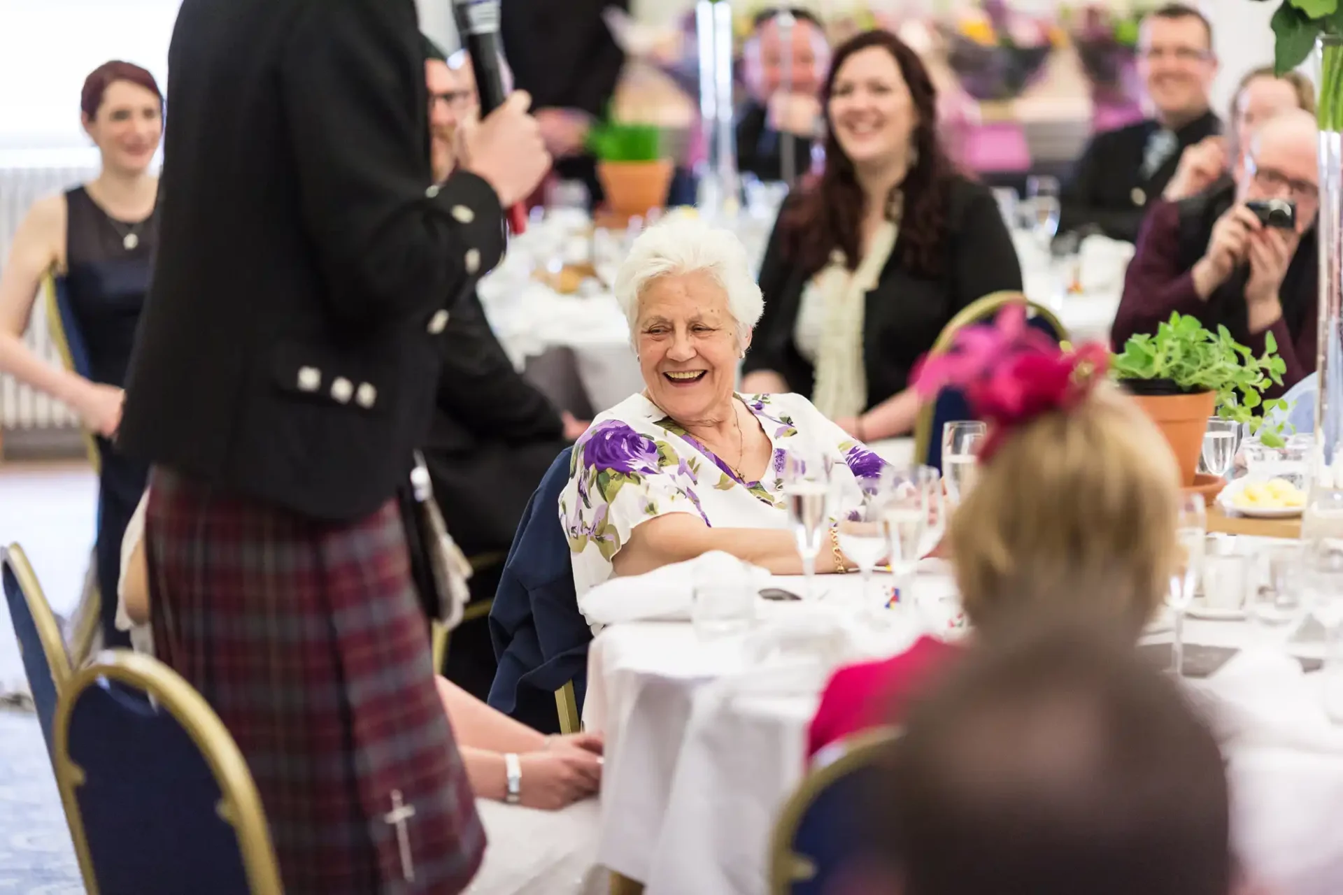 Elderly woman in a floral dress laughing joyfully at a table during a festive event with guests and a waiter in a kilt.