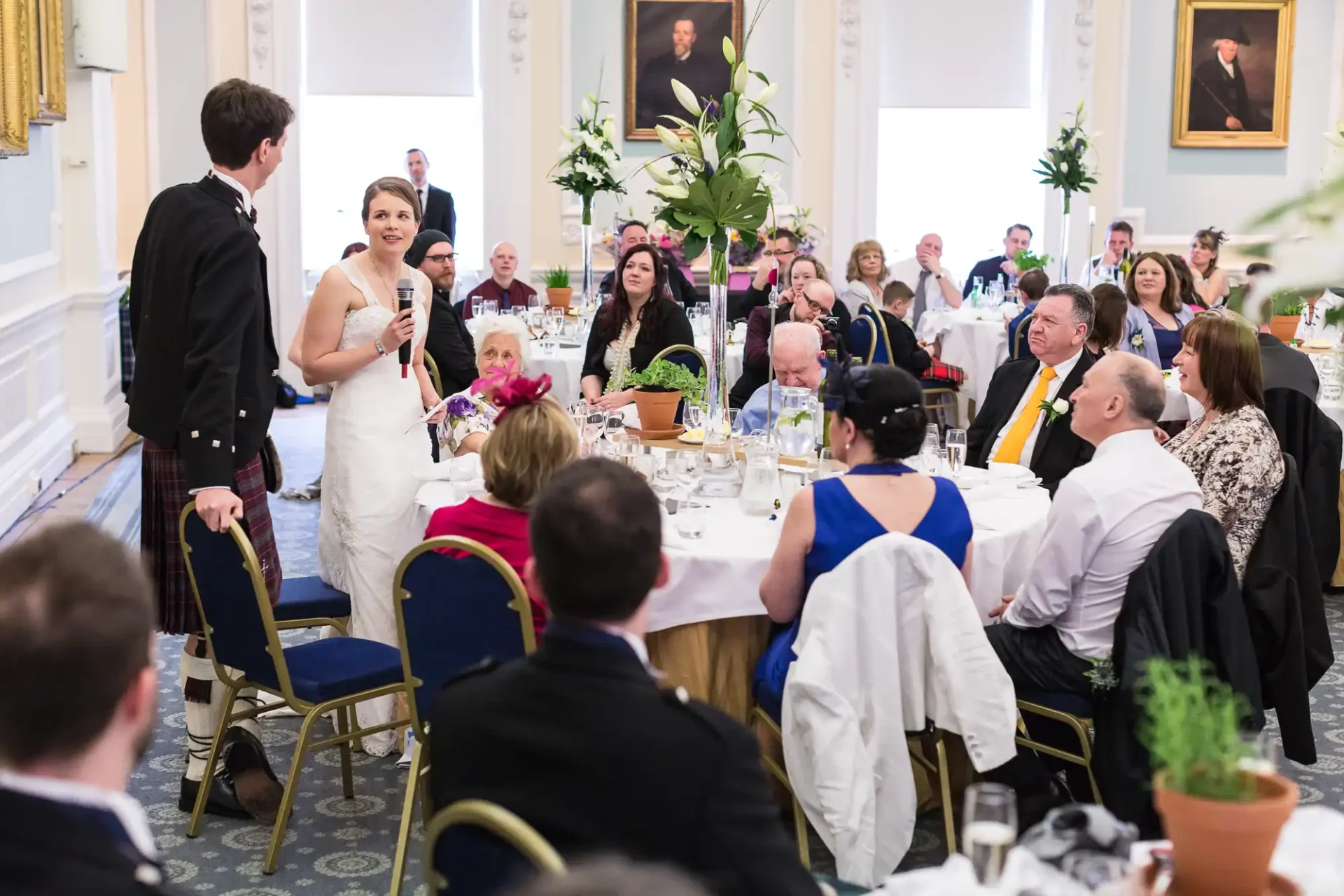 A bride and groom stand at their wedding reception, engaging with seated guests in an elegant dining hall.