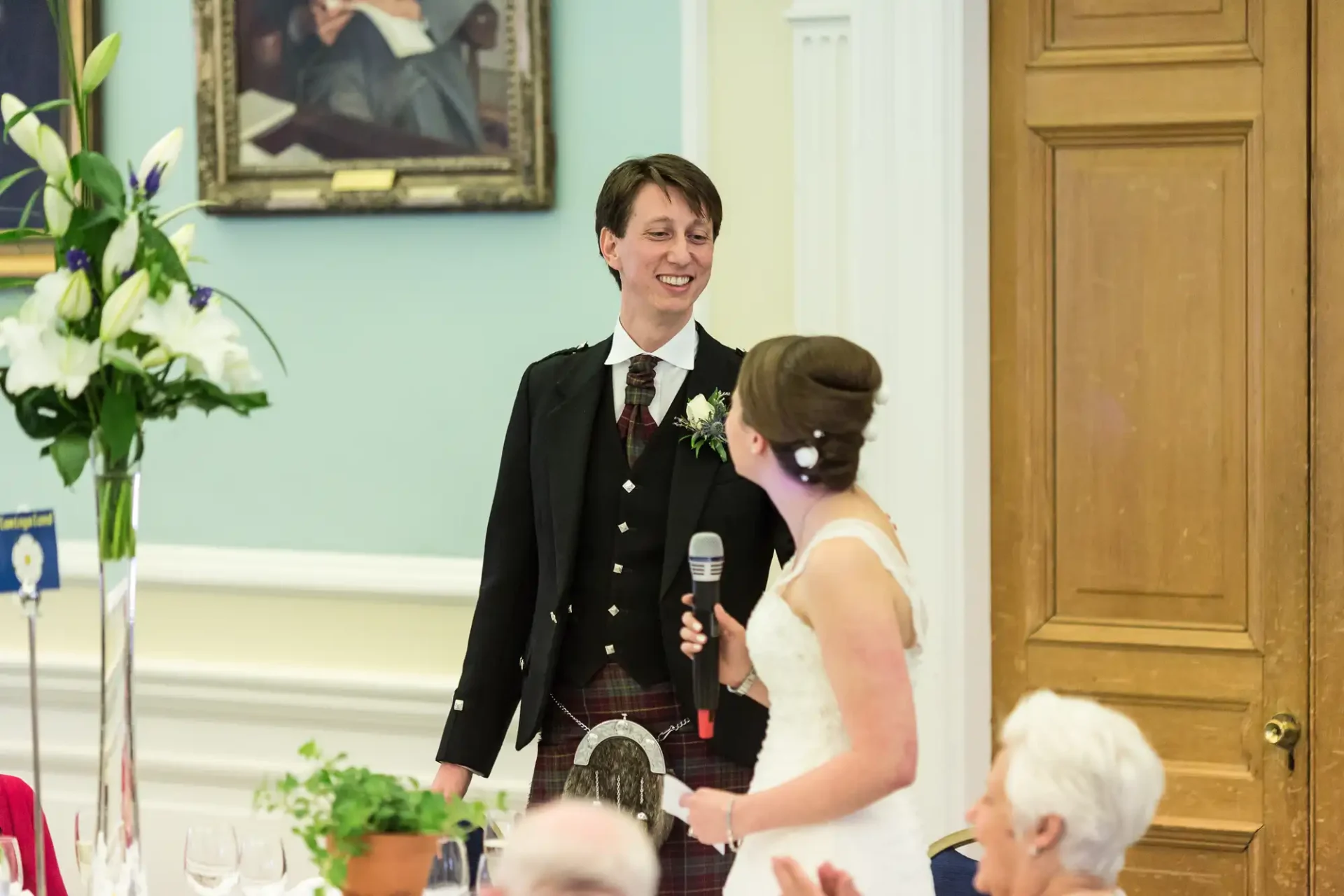 A person in a kilt and jacket smiling at a bride speaking into a microphone at a wedding reception.