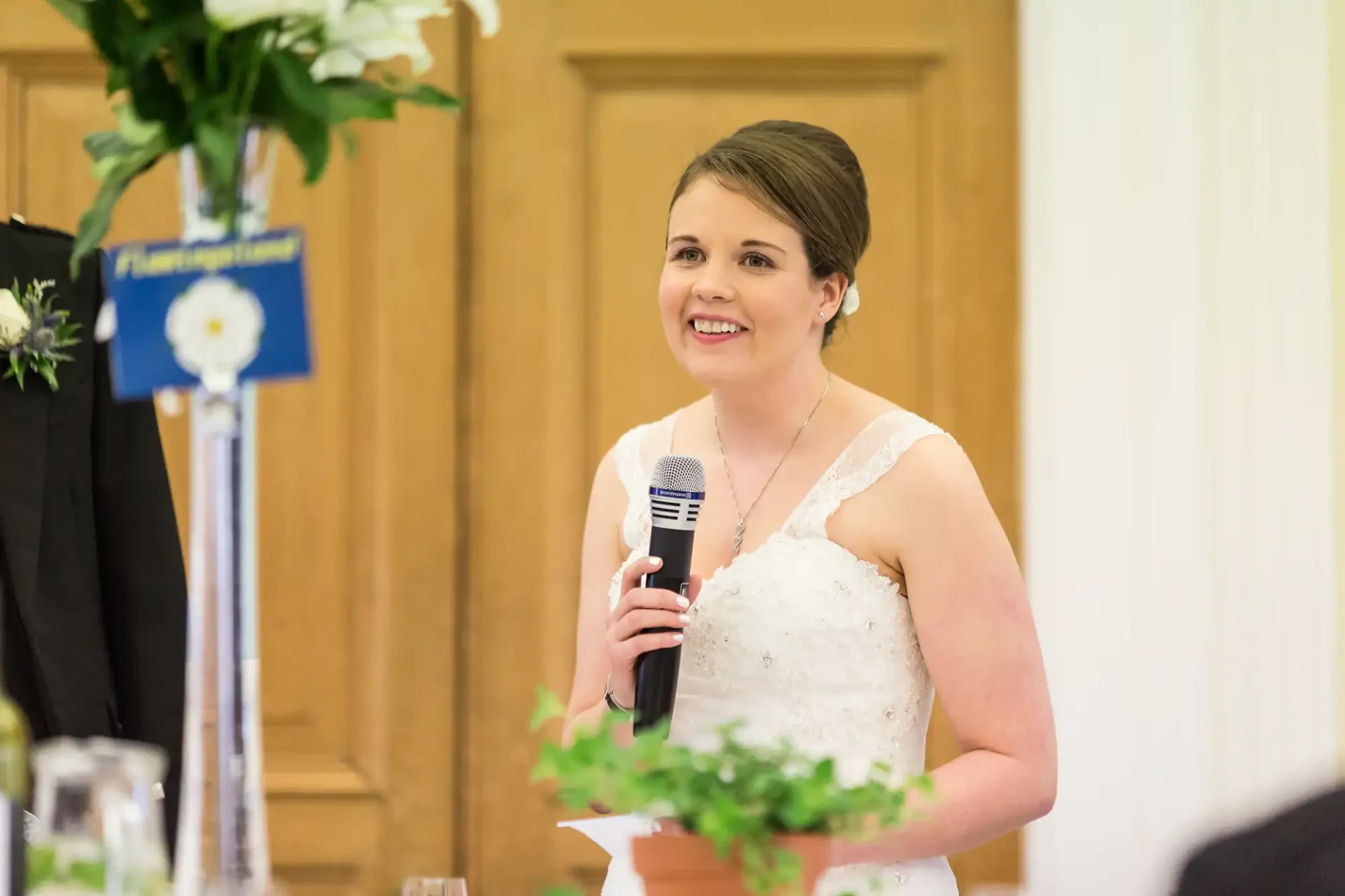 A bride in a white dress speaking into a microphone at a wedding reception, smiling, with guests and floral decorations in the background.