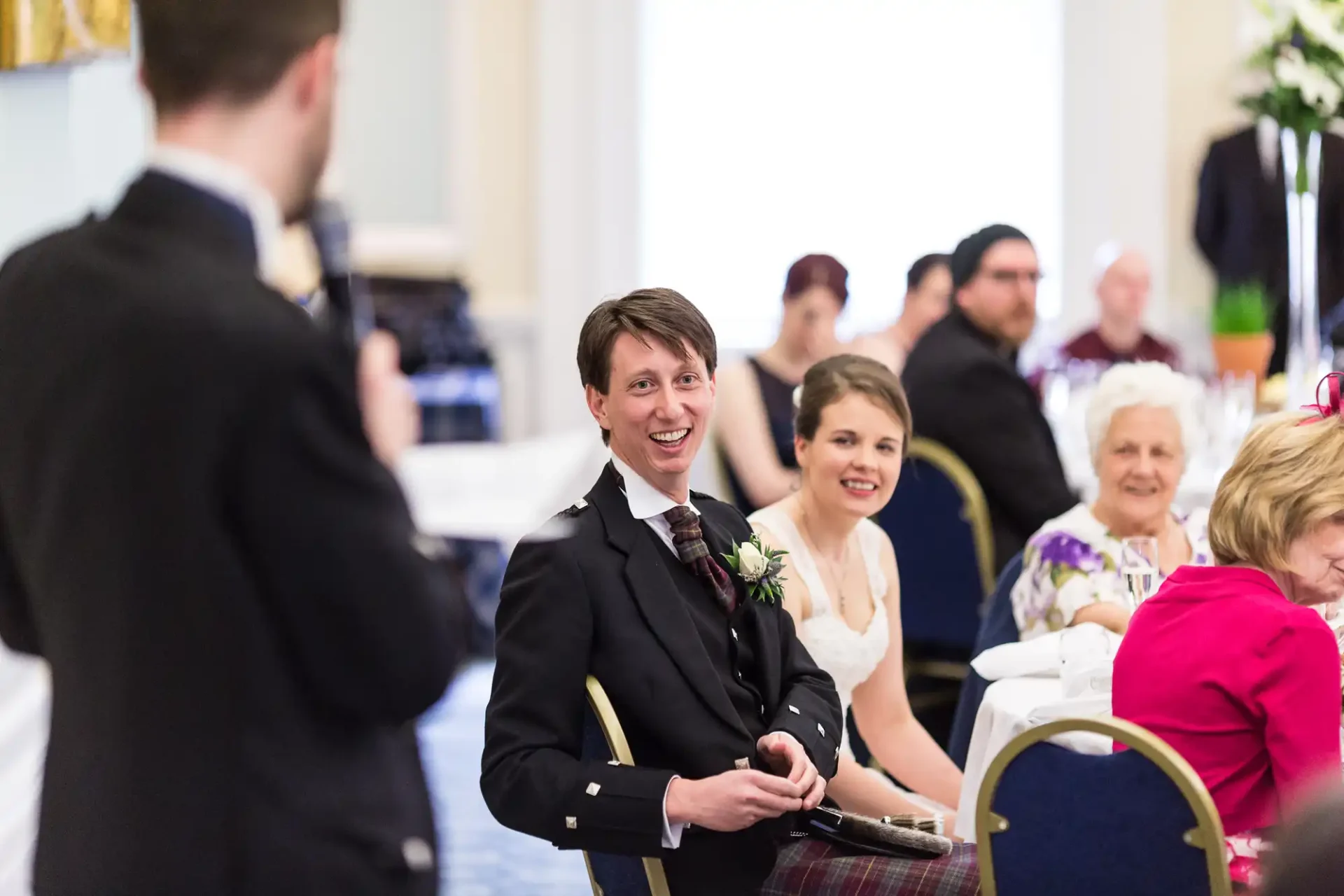 A smiling groom in a suit with a boutonniere holds a microphone, seated next to a bride in a hall filled with guests.