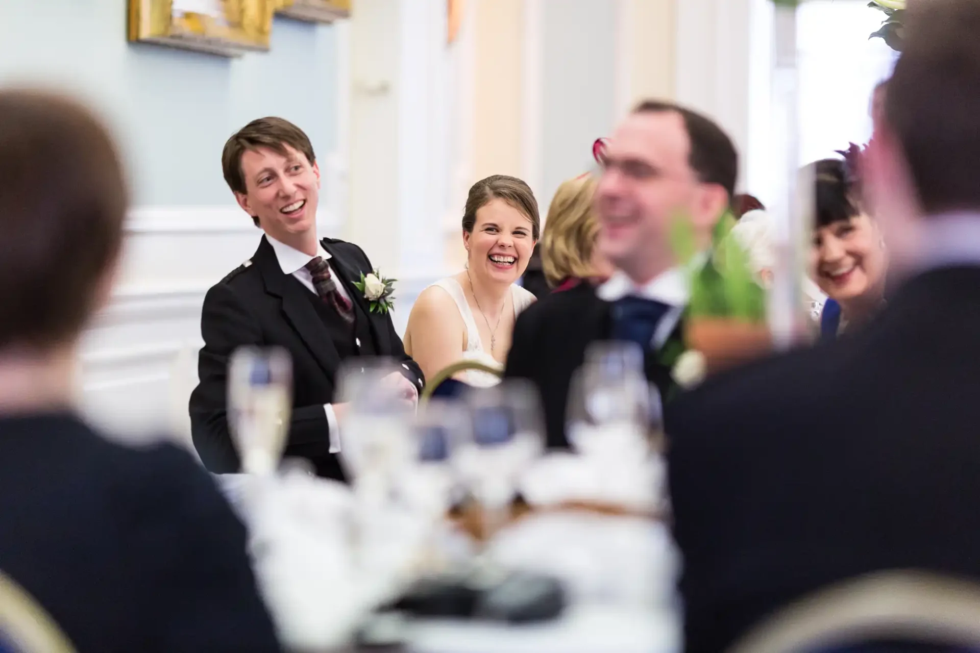 A bride and groom laugh joyously at a wedding reception table surrounded by guests in a well-lit room.