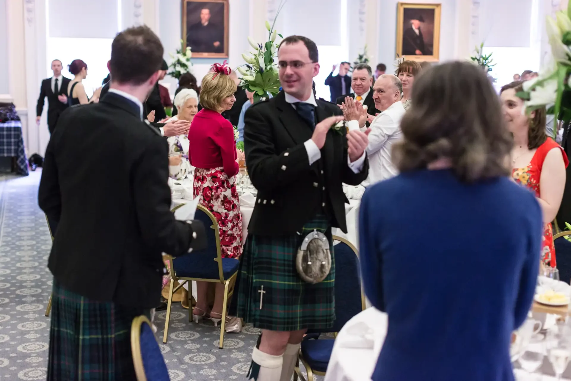 Men in kilts clapping at a lively indoor event with seated and standing guests, some engaged in conversation.