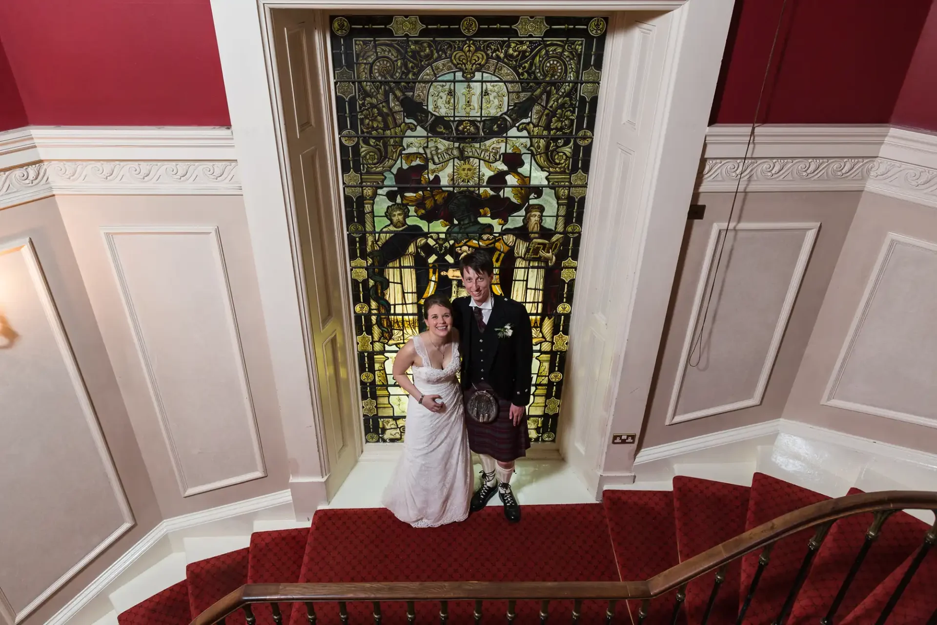 A bride in a white dress and a groom in a kilt standing on a staircase in front of a large, ornate stained glass window.
