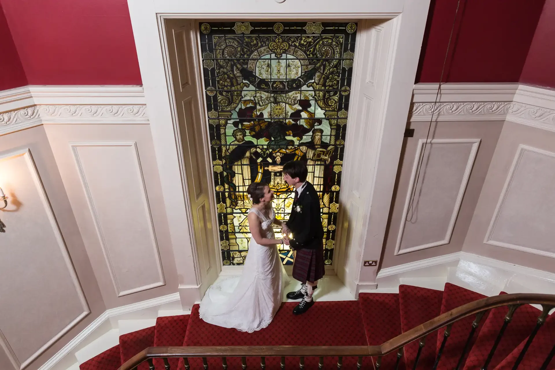 Bride in white dress and groom in kilt standing at the bottom of a grand staircase beneath an elaborate stained glass window, inside an elegant hall.