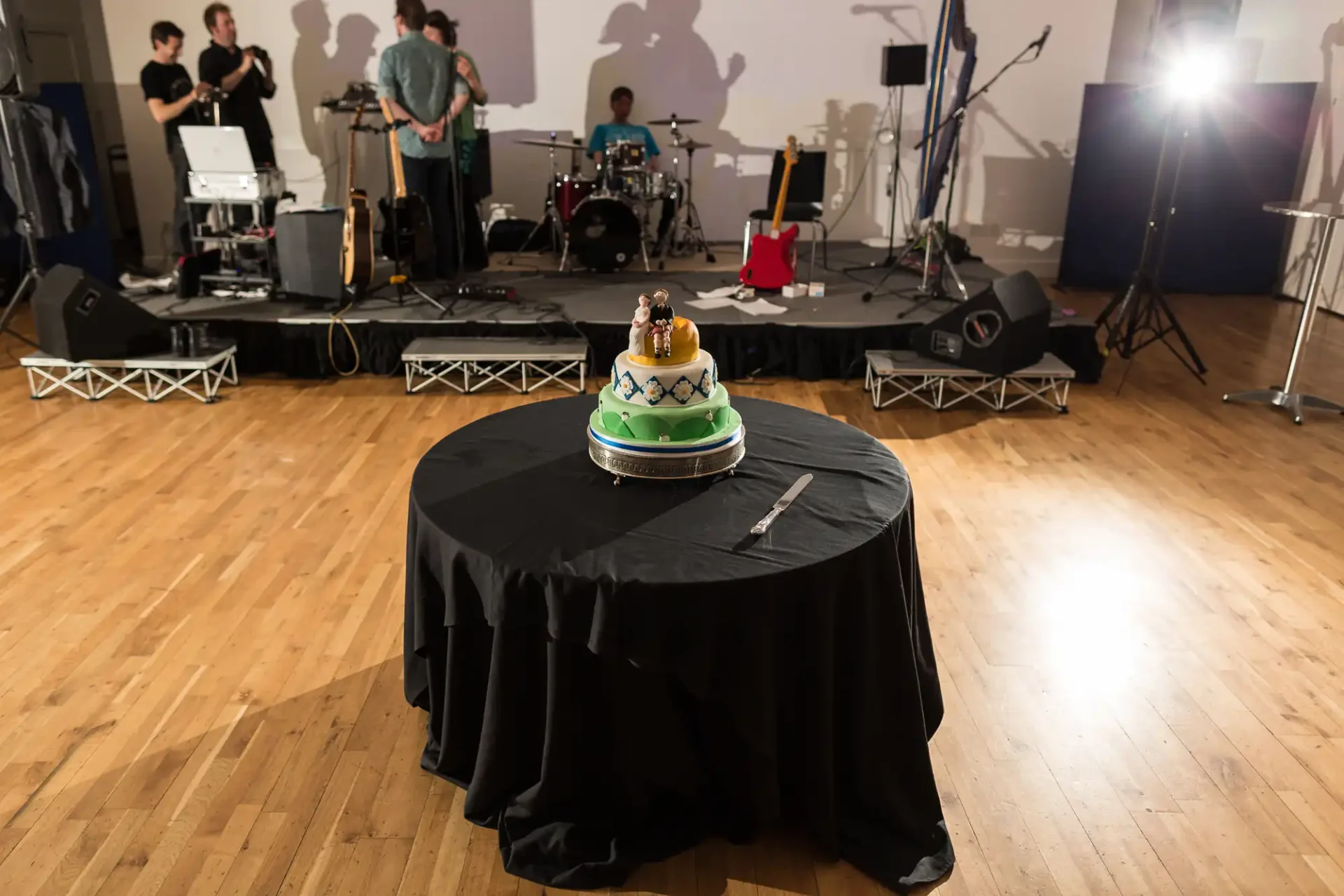 A wedding cake with a same-sex couple topper on a table, with a band performing in the background on a stage.