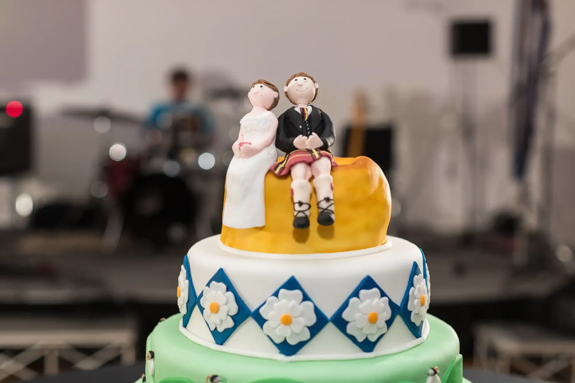 Wedding cake topped with a figurine of a bride and groom sitting on a bench, with a colorful floral design on the tiers, set against a blurred background of a party.