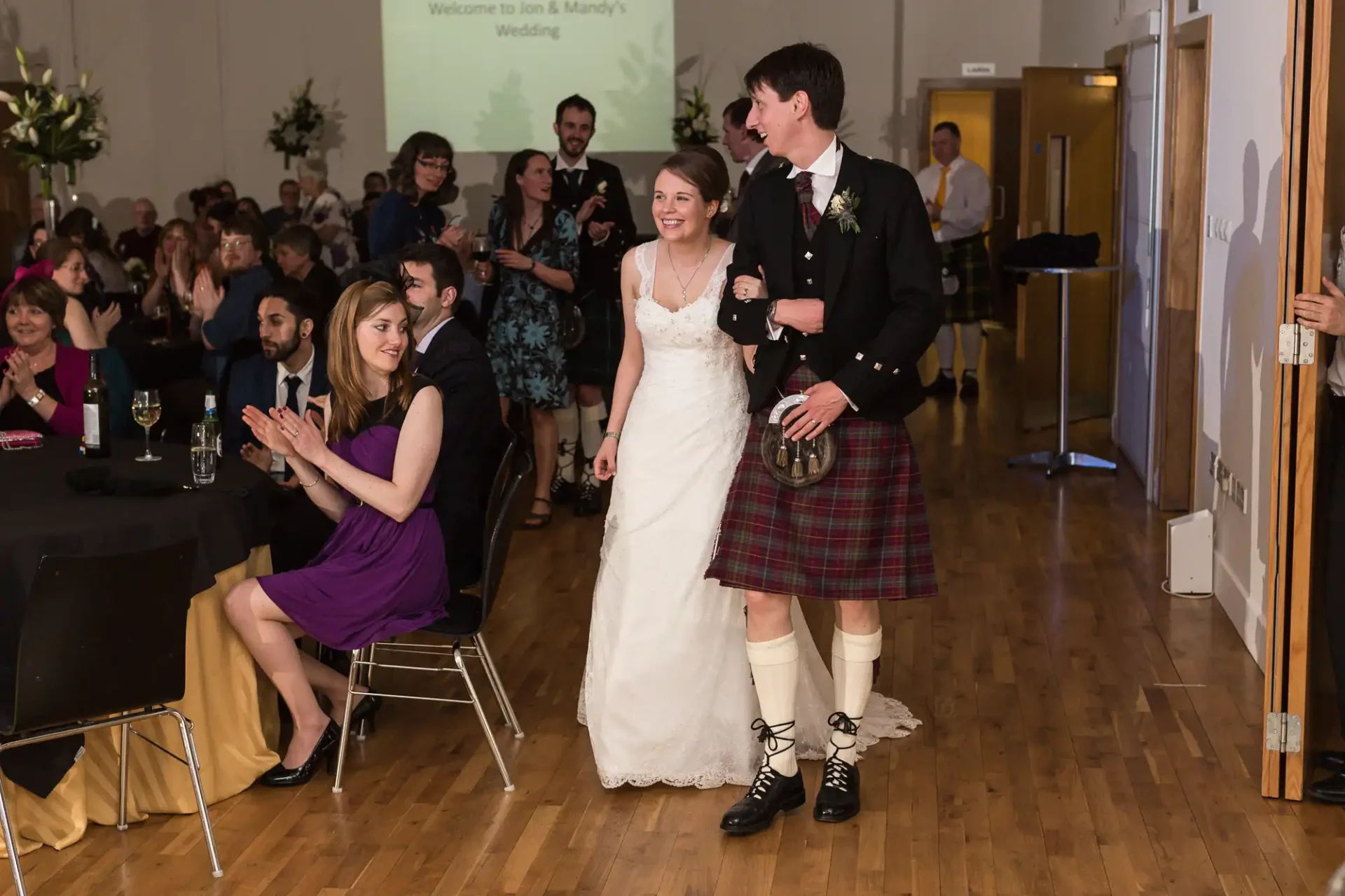 A bride and groom in traditional scottish attire smiling and walking through a hall as guests applaud at their wedding reception.