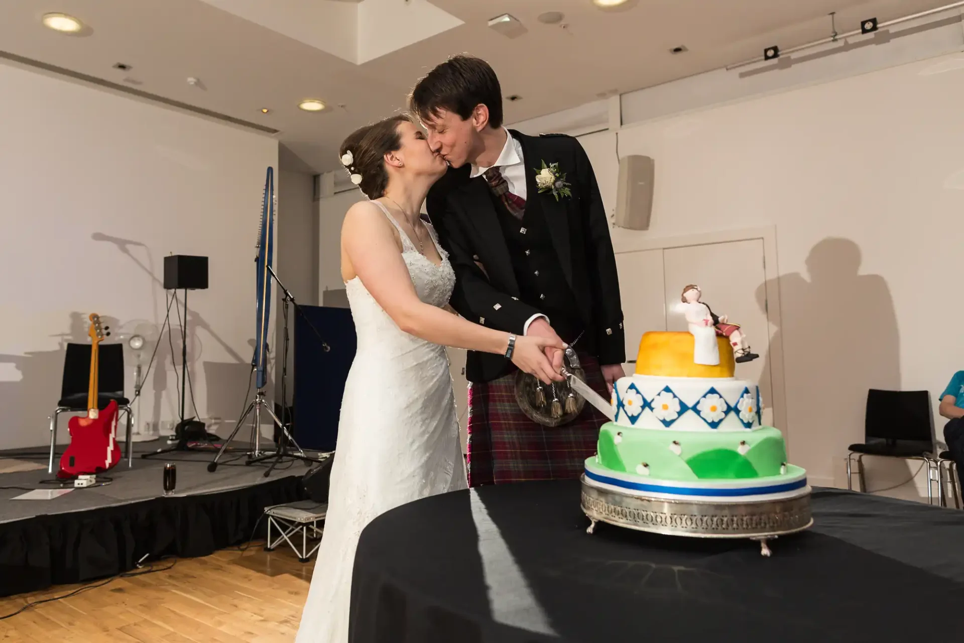 A bride and groom in wedding attire kiss as they cut a three-tiered cake at their reception. the groom wears a kilt and the cake is decorated in bright colors.