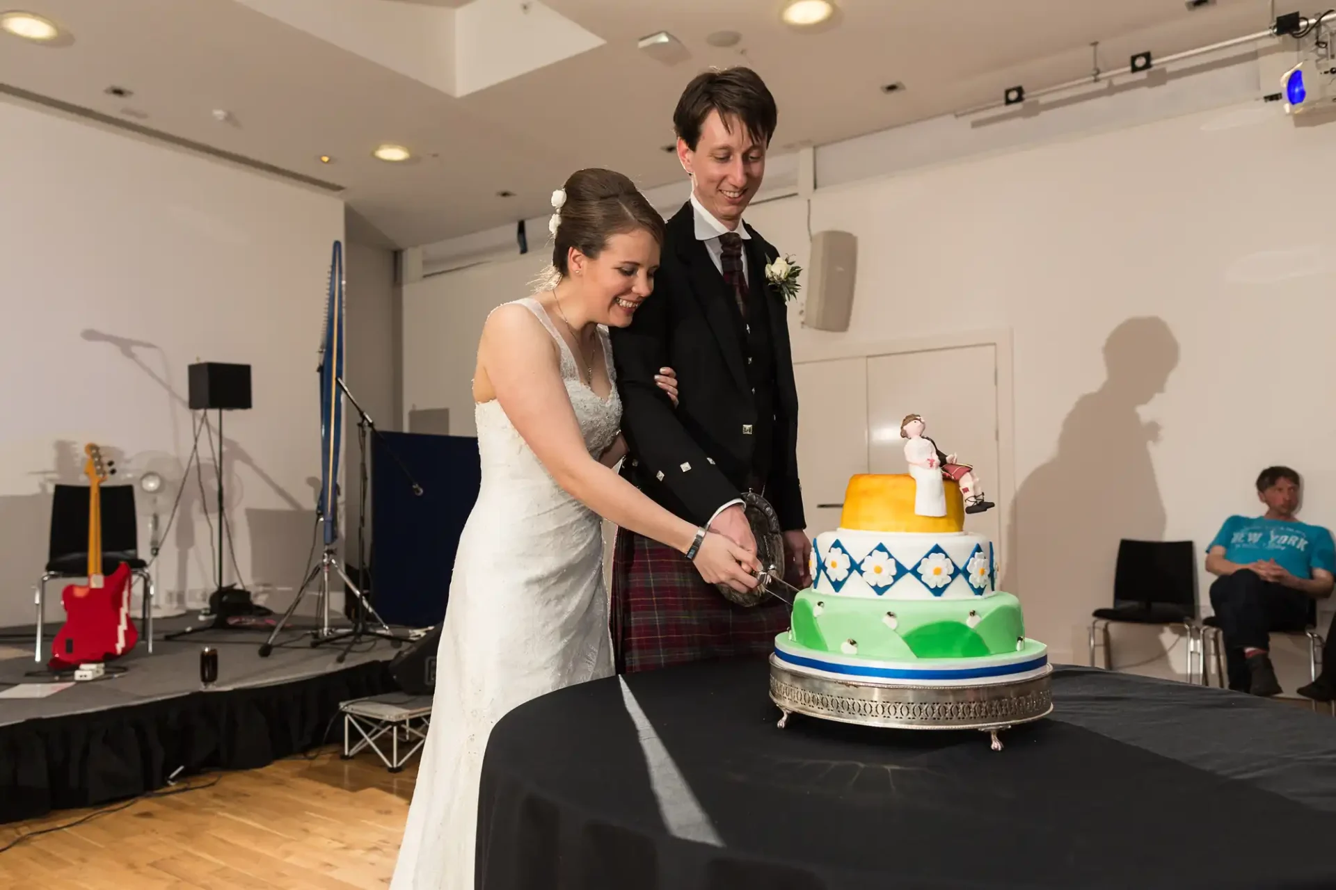 A bride and groom in formal attire cutting a three-tiered, colorful wedding cake in a room with a stage and musical equipment in the background.