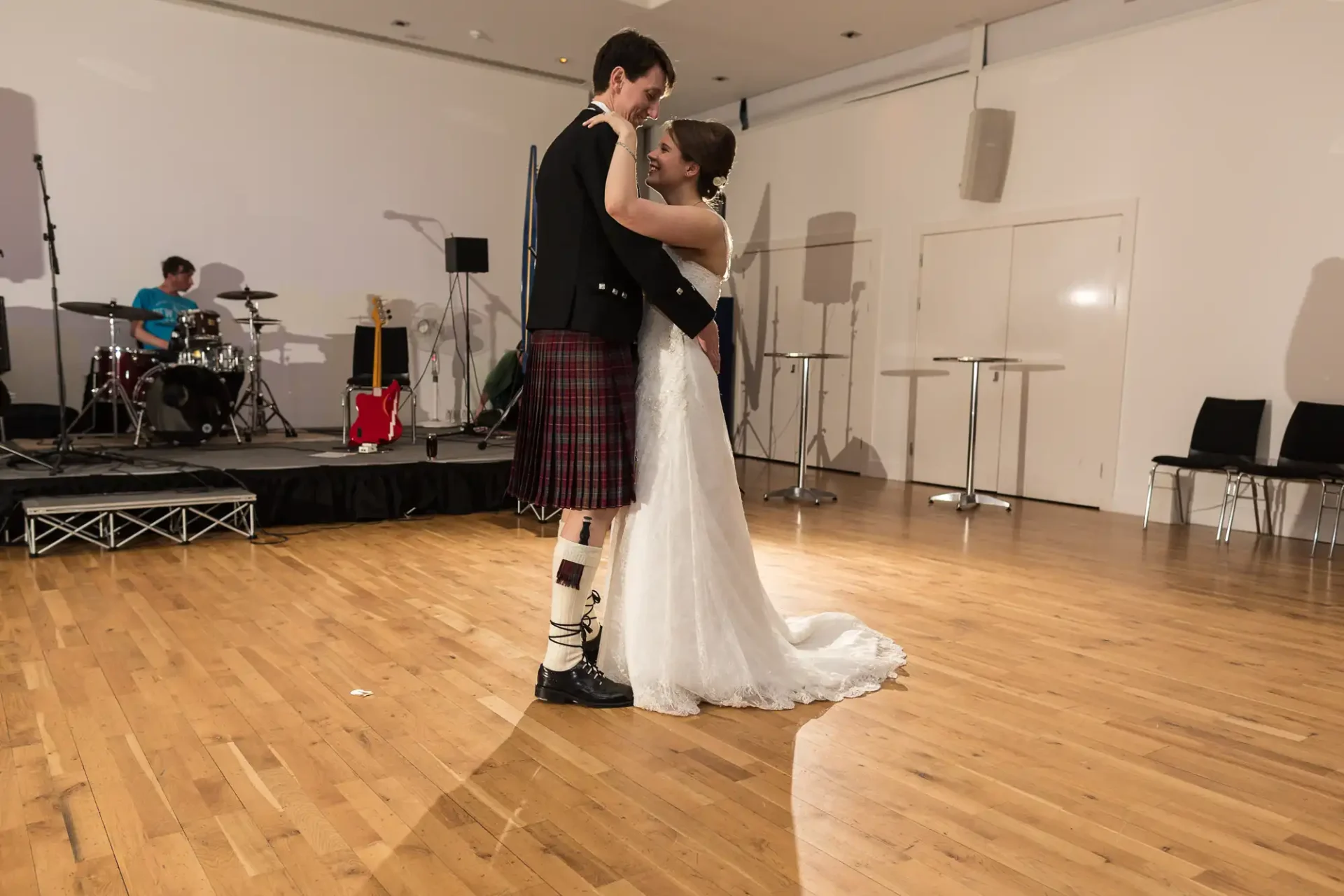 A bride and groom in a dance pose during their wedding reception; the groom is wearing a kilt, and a drummer is visible in the background.