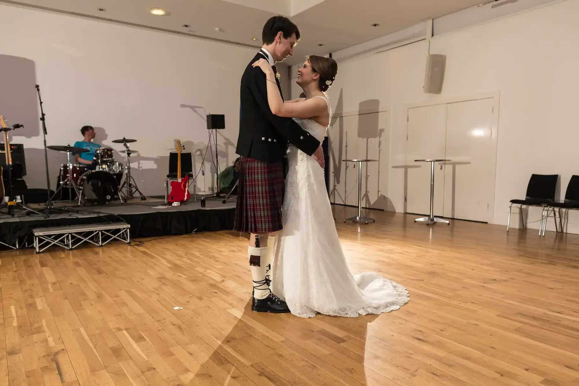 A bride in a white dress and a groom in a kilt share a romantic dance at their wedding reception, with a band playing in the background.