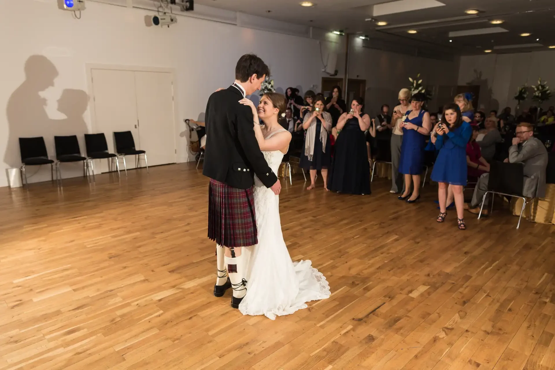 A bride and groom share their first dance at a wedding reception, surrounded by guests watching and taking photos. the groom wears a kilt and the bride is in a white dress.