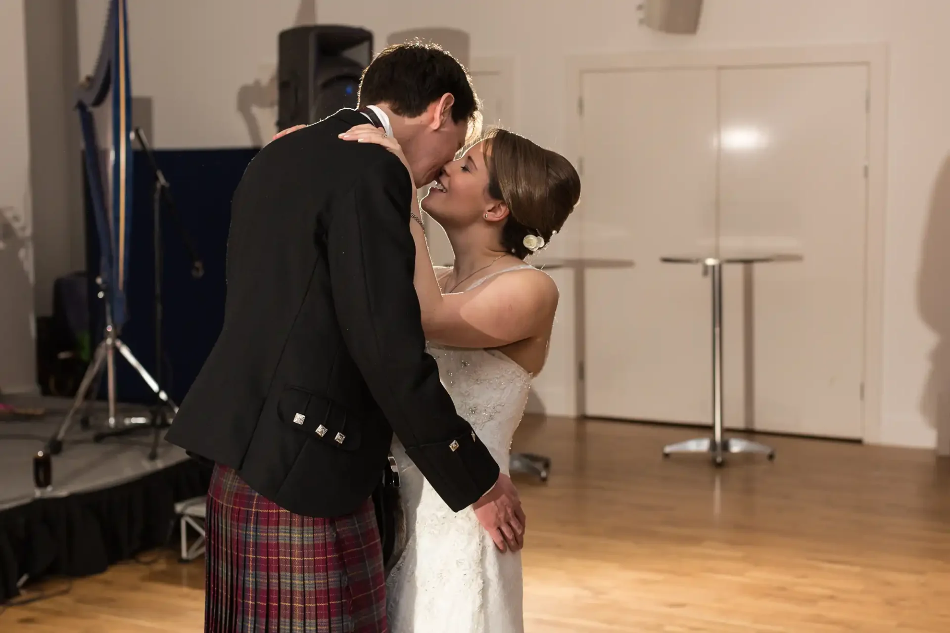 A bride and groom sharing a kiss on their wedding day, with the groom wearing a kilt and the bride in a white gown, in a room with musical equipment in the background.