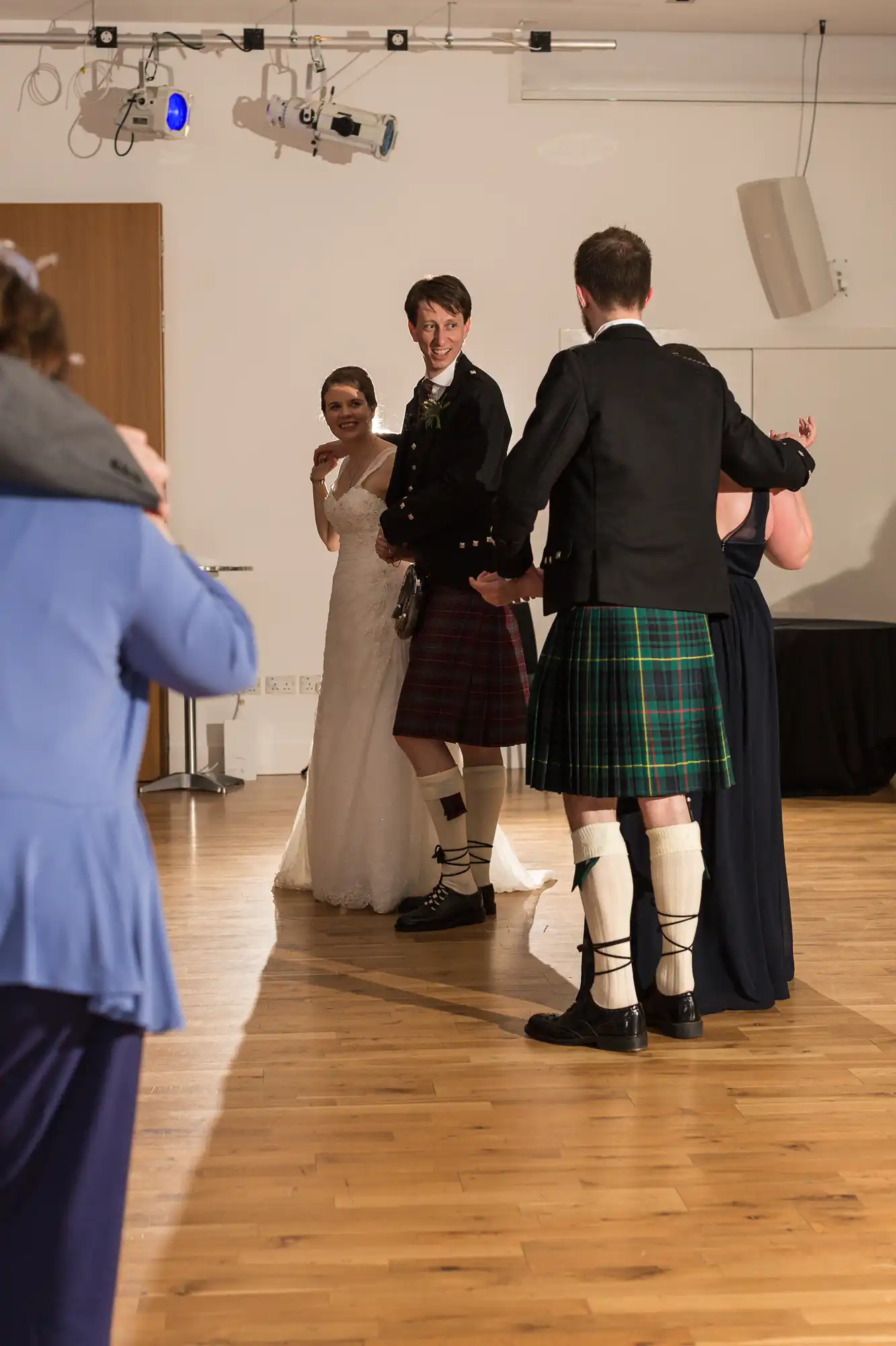 A bride and groom smile as they participate in a dance, surrounded by guests in a well-lit hall, with traditional scottish kilts visible.