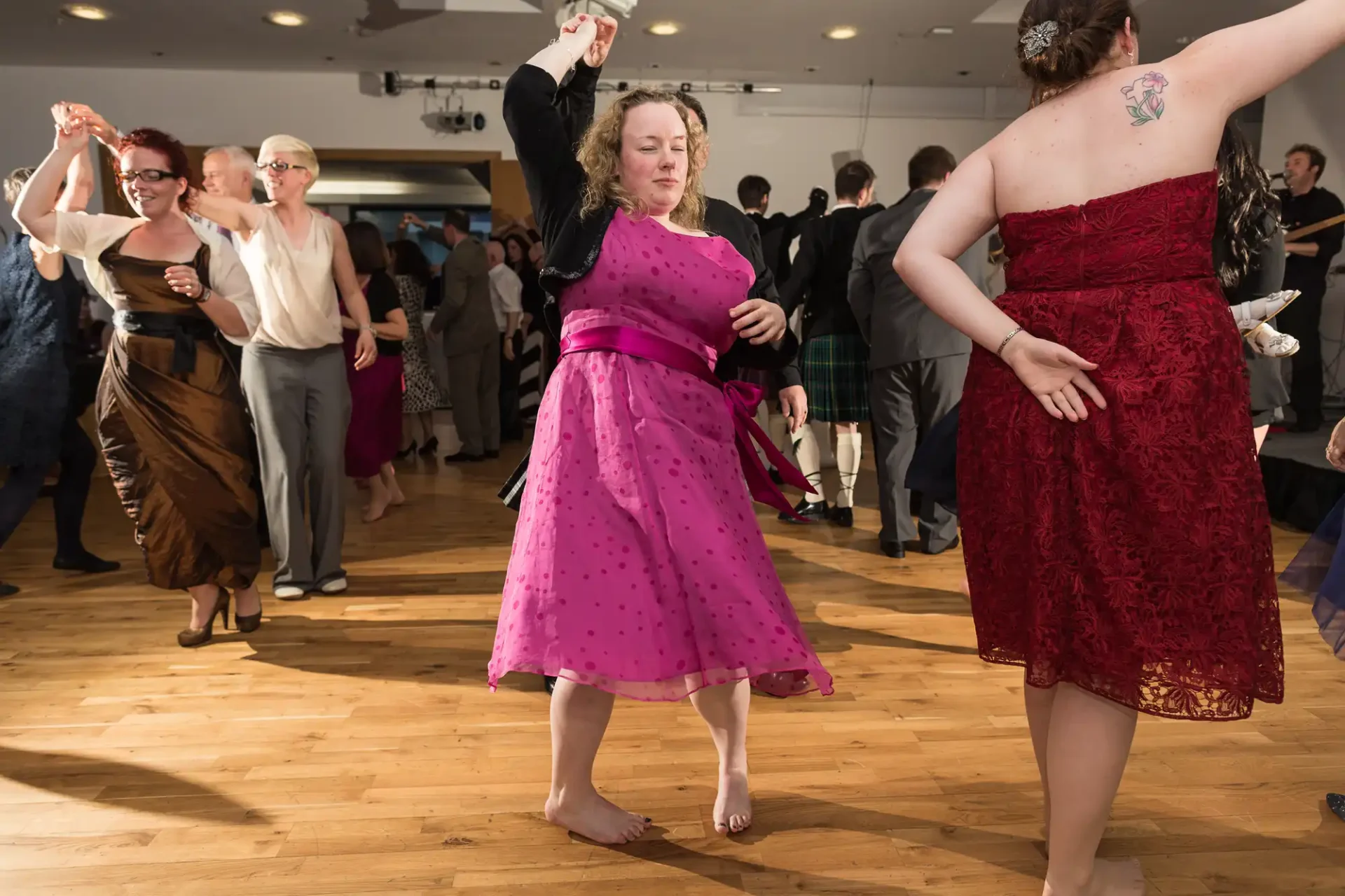 A group of people joyfully dancing at a formal event, with one woman in a pink polka-dot dress featured prominently in the foreground.