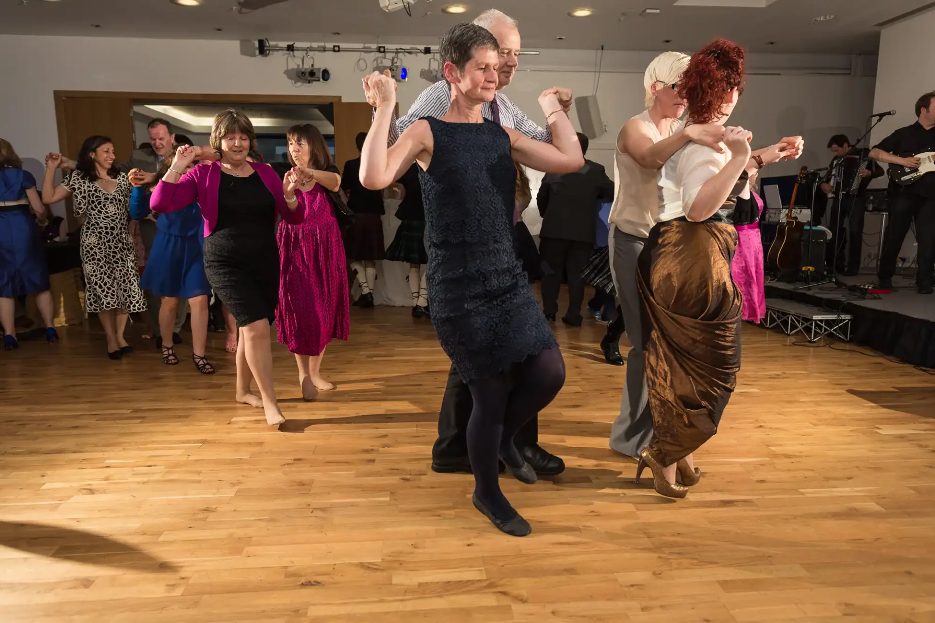 Adults enjoying a lively dance at a party in a ballroom with a wooden floor, as a band plays in the background.