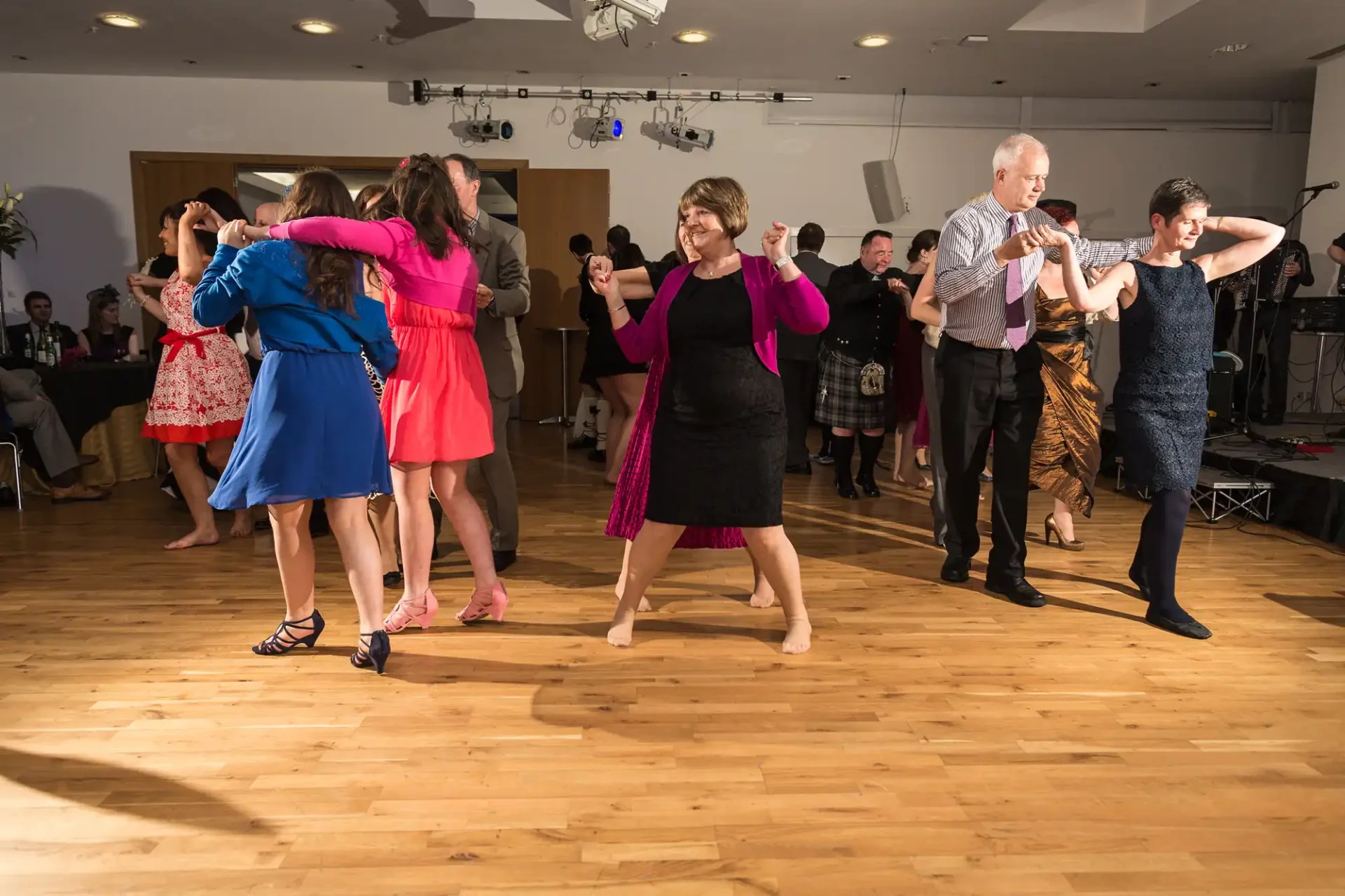 People dancing in pairs at a lively indoor event, on a wooden floor with a band performing in the background.