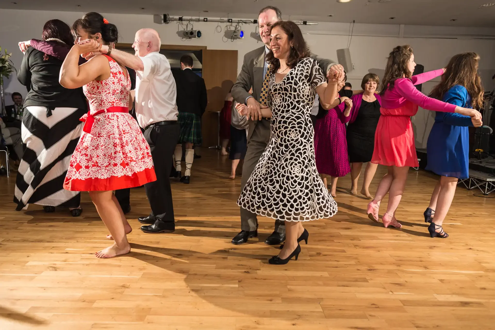 People of various ages joyfully dancing in pairs at an indoor event with a wooden floor.