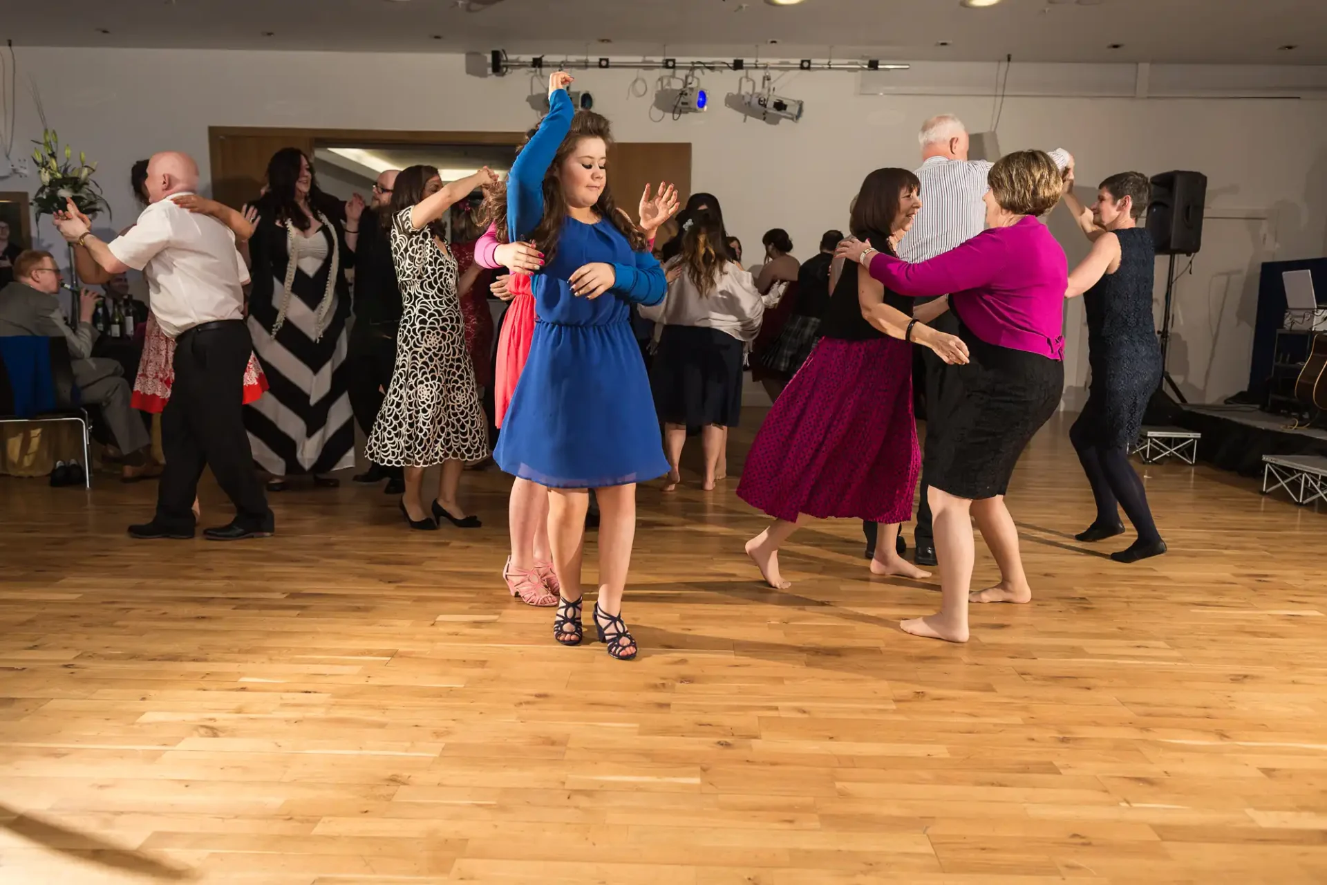 A young woman in a blue dress dances alone with a red ribbon at a lively party while other couples dance around her.